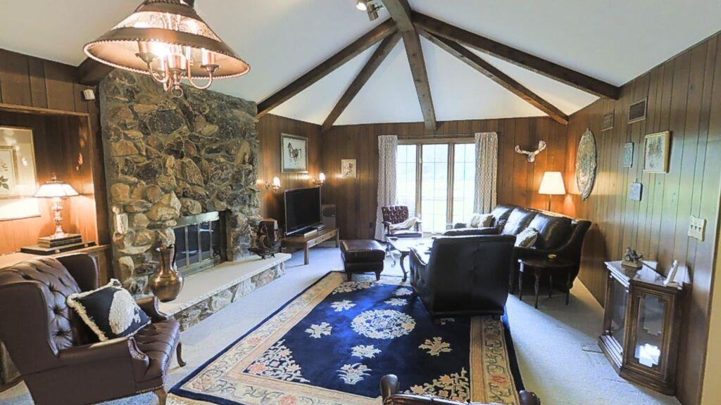 Cozy living room with a stone fireplace, wooden beams, leather furniture, stylish decor, large window, elegant rug, and warm lighting.