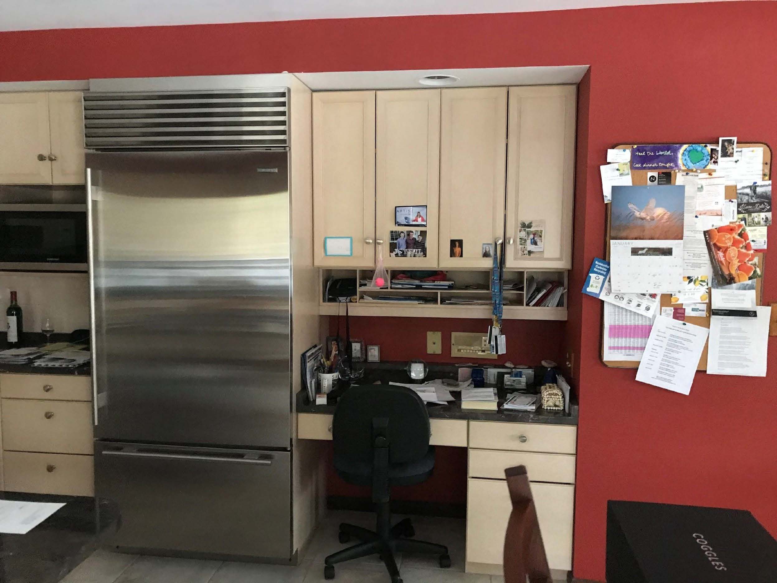A kitchen corner with a red wall, stainless steel refrigerator, desk area, office chair, and a bulletin board filled with papers and photos.