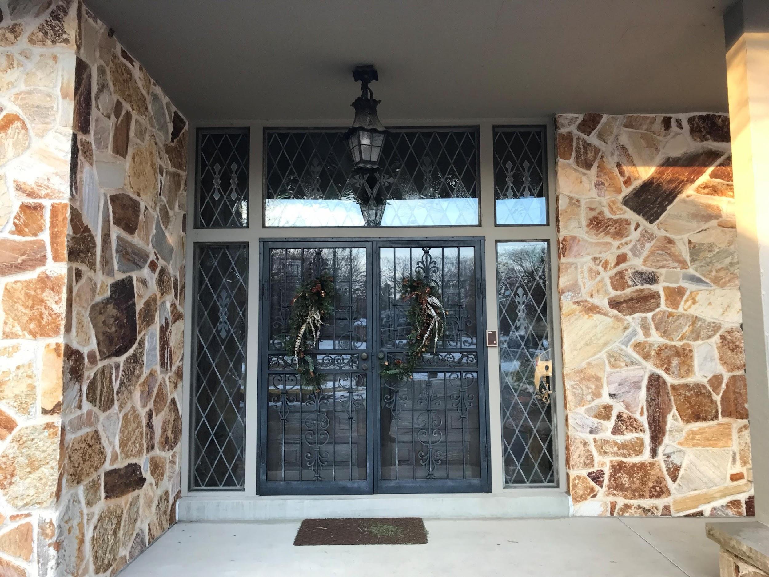 Elegant entrance with decorative metal door, stone walls, and diamond-patterned windows. Wreaths hang on doors under an elaborate lantern overhead.