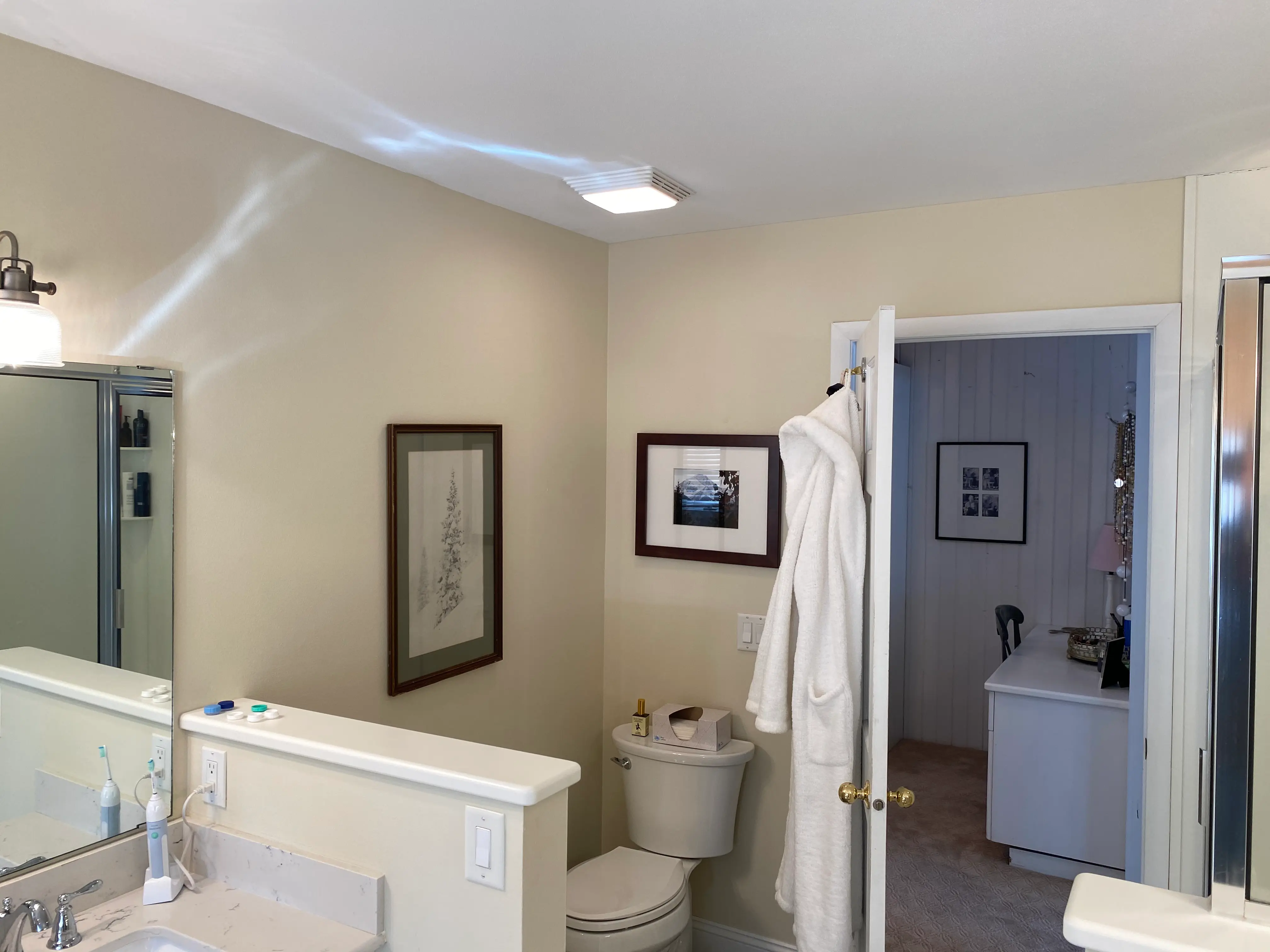 Bathroom with a white robe on the door, framed art, sink, and open door revealing a glimpse of another room.