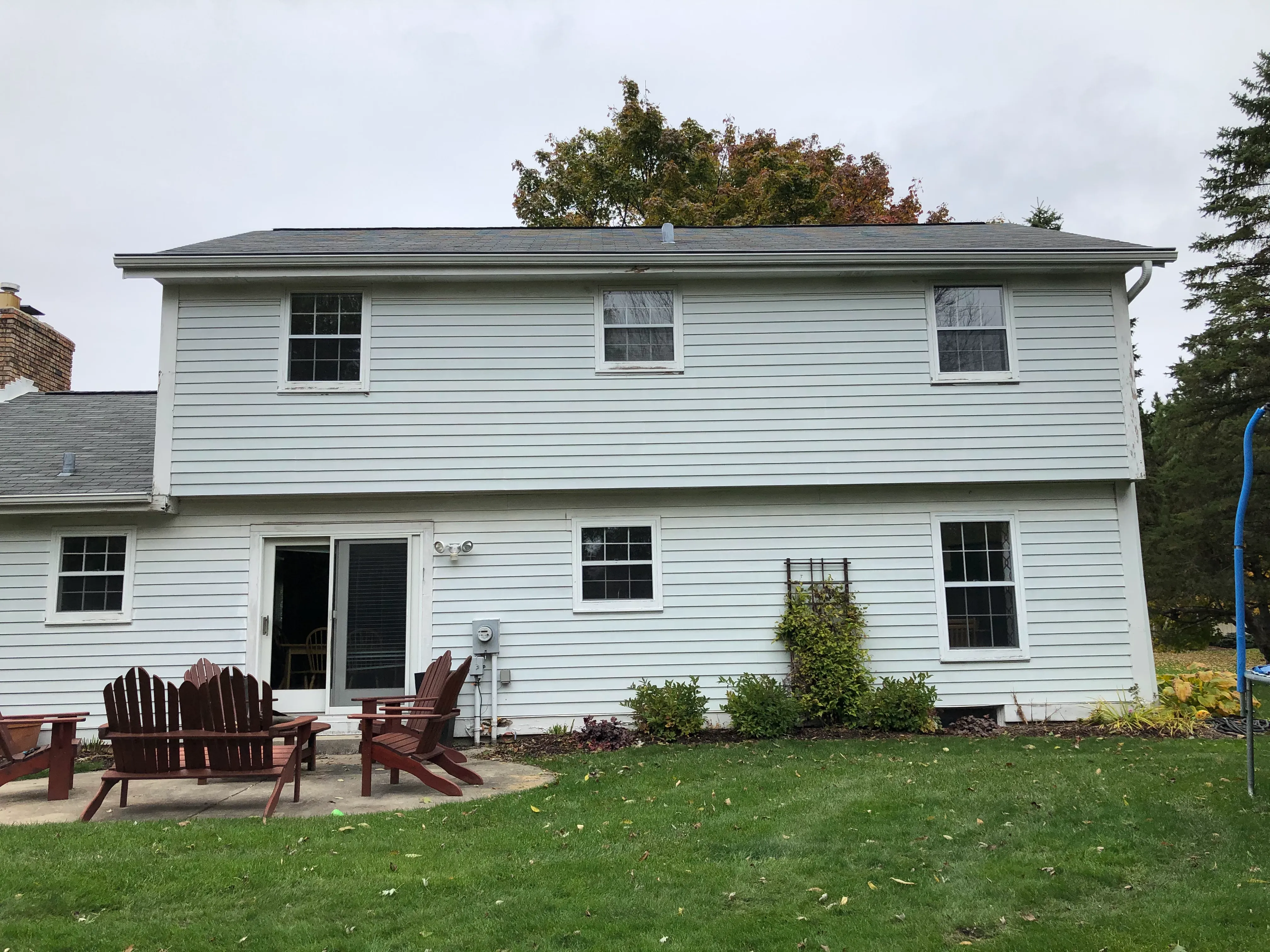 A white two-story house with wooden chairs on a patio. The yard is green with trees in the background.