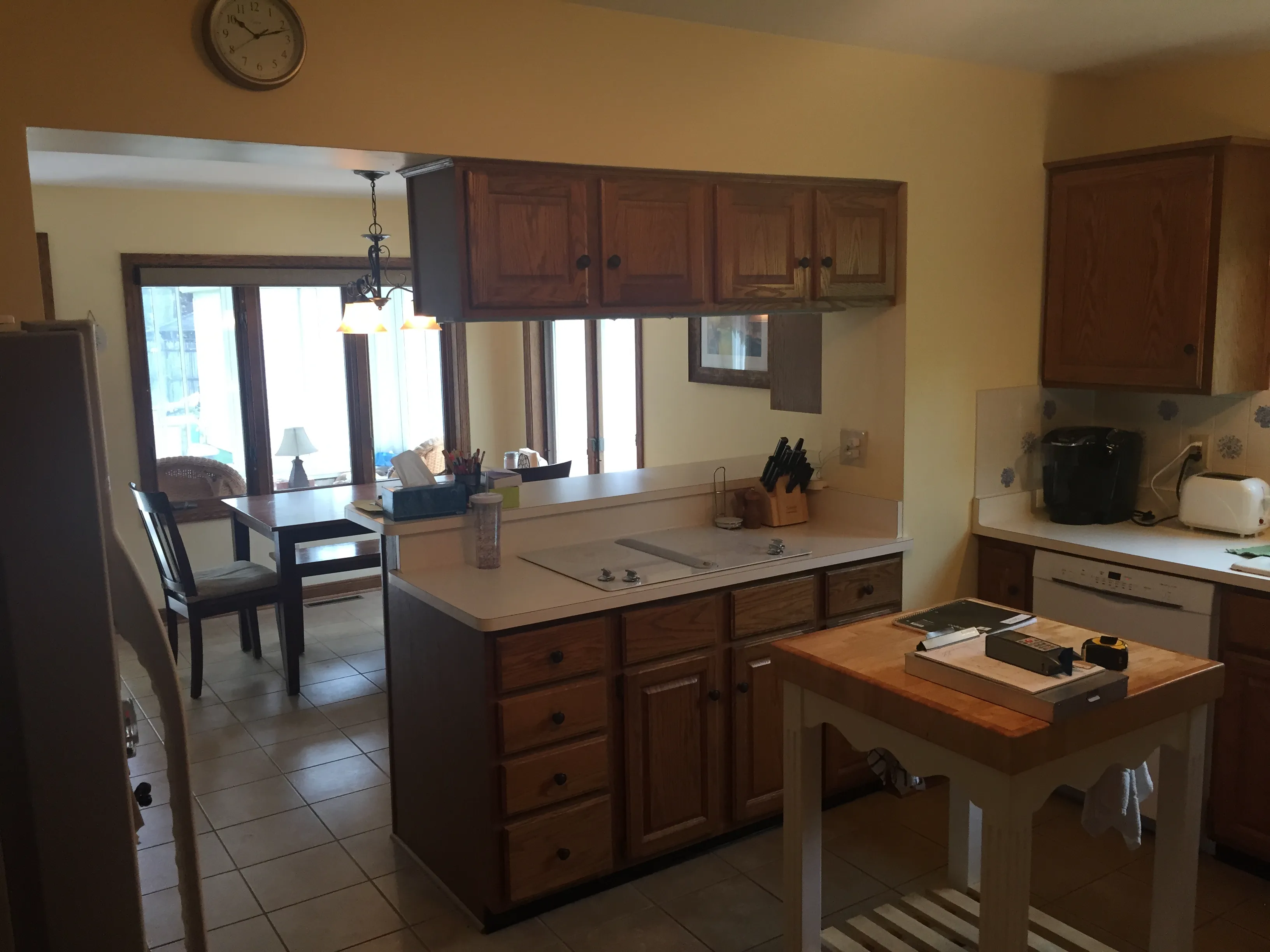 A cozy kitchen with wooden cabinets, island, clock, and adjacent dining area featuring large windows, wooden table, and chairs. Bright, inviting space.