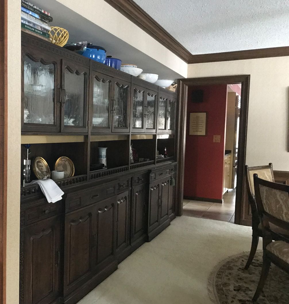 Ornate wooden cabinet with glass doors in a cozy dining area, featuring decorative plates, books, and bowls on the countertop and upper shelf.