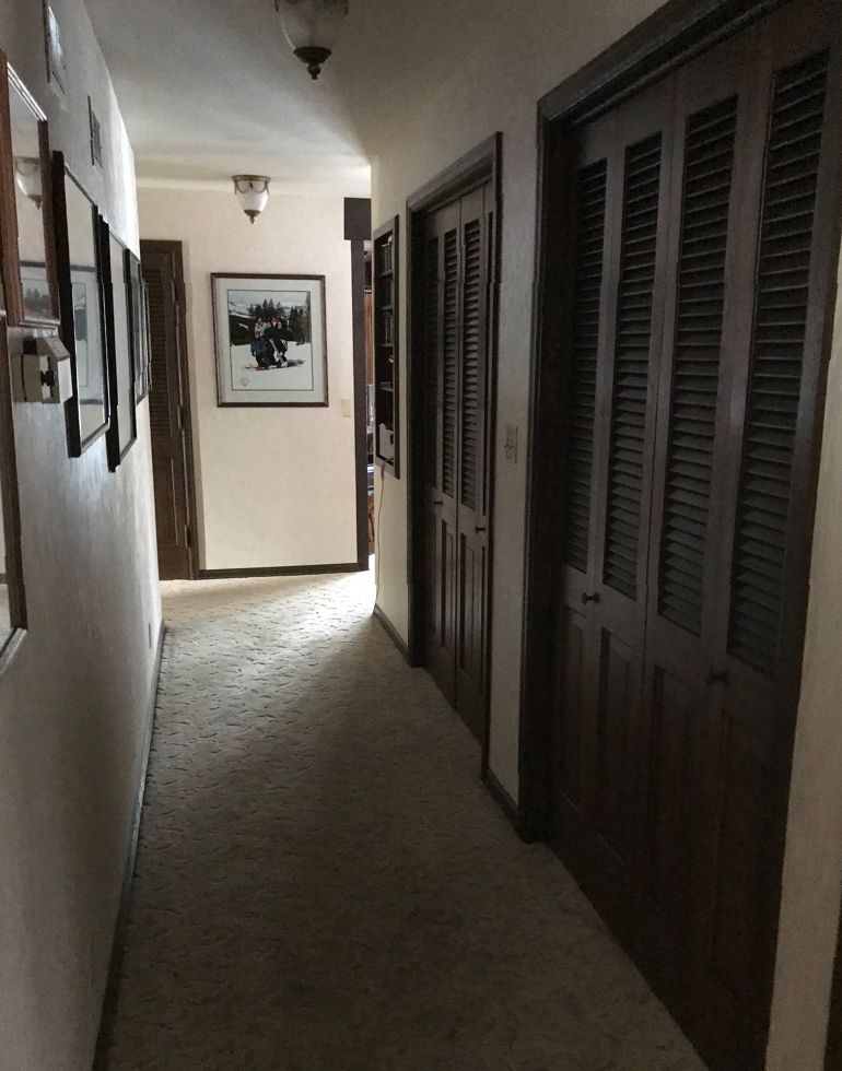 A dimly lit hallway with carpet, framed pictures, wooden doors, and ceiling light fixtures leading towards a partially open room.