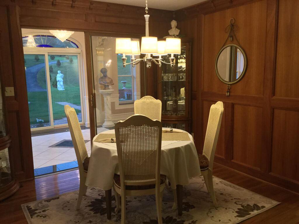 Wood-paneled dining room with round table, statues, and glass cabinet. Large window reveals garden view with statues outside. Elegant ambiance.