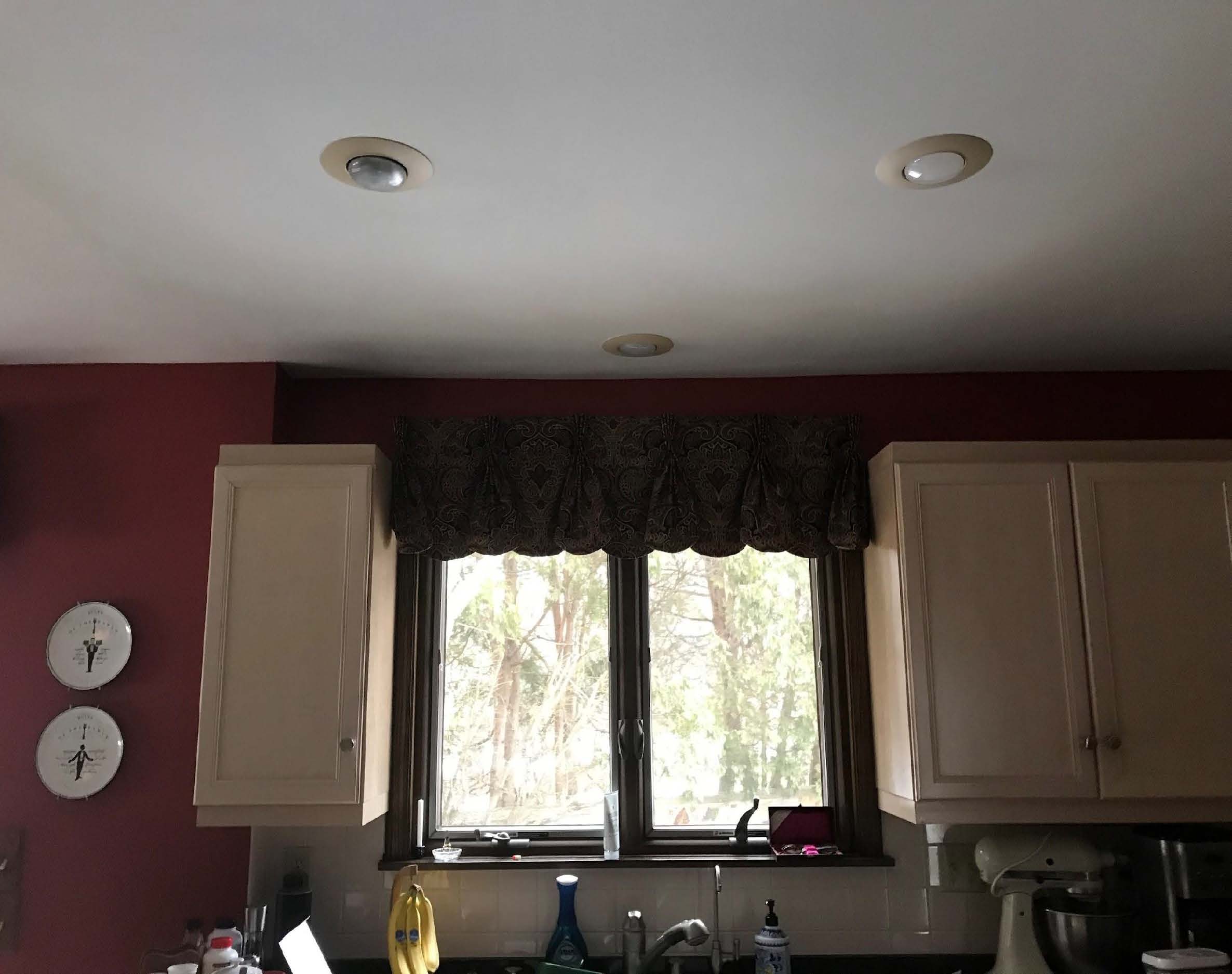 A kitchen with maroon walls features wooden cabinets, a central window, ceiling lights, a mixer, plates on the wall, and various items.