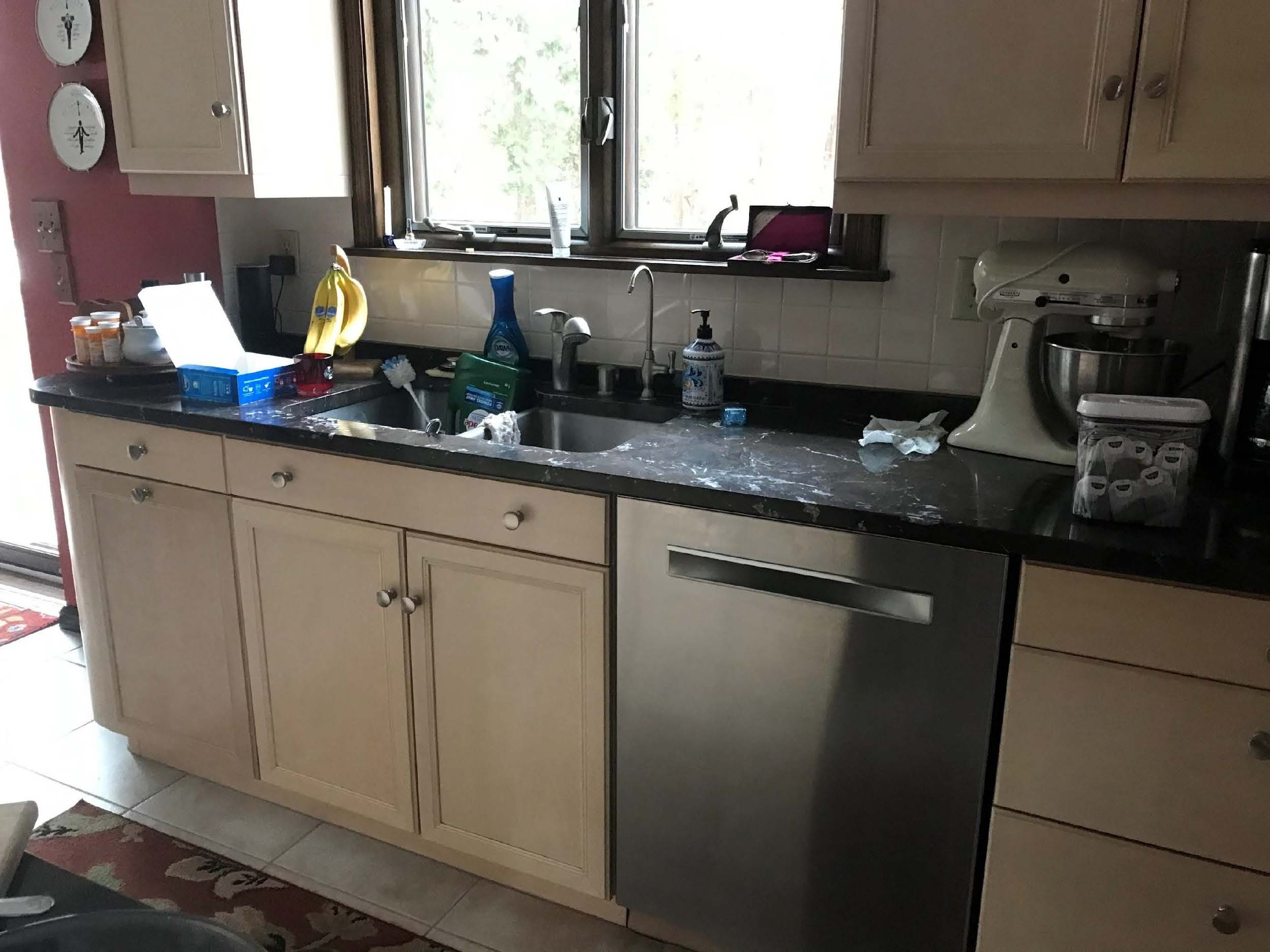 A kitchen scene with cabinets, a black countertop, mixer, bananas, and various cleaning supplies. A window provides natural light above the sink.