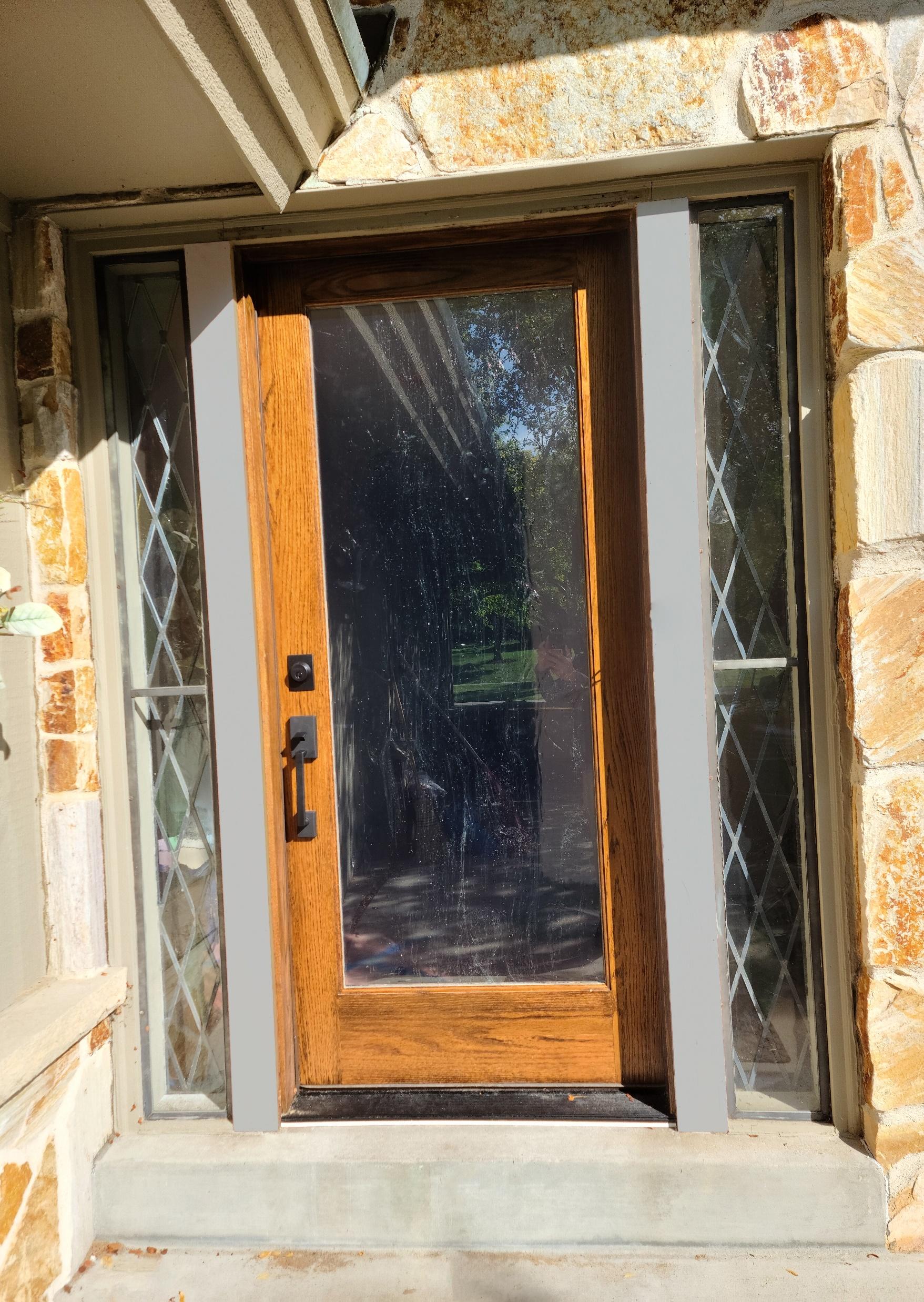 A wooden door with diamond-patterned glass sidelights, set in a stone facade, reflects outdoor greenery under bright sunlight.
