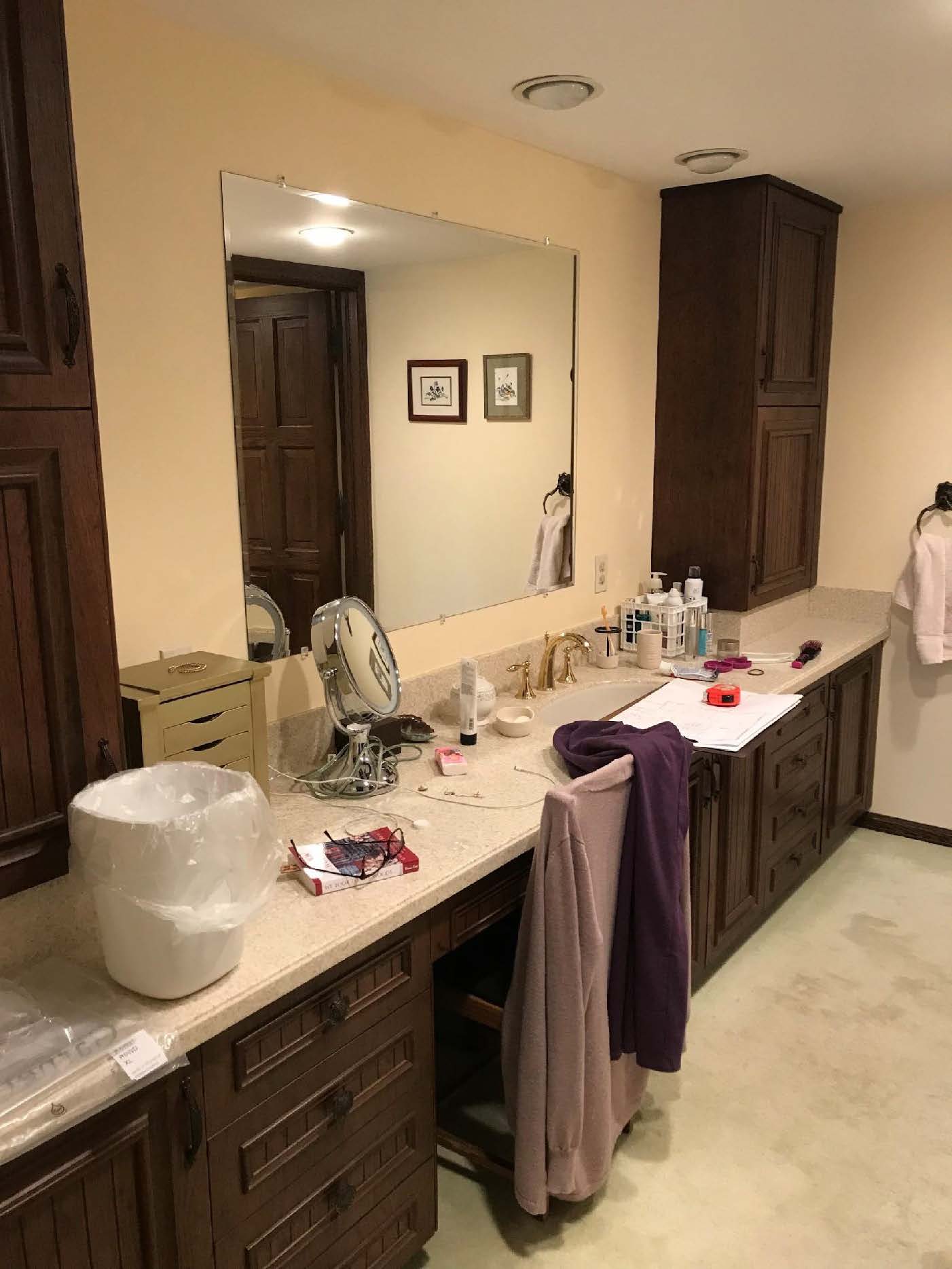 A bathroom with a large mirror, wooden cabinets, and various toiletries and clothing items scattered on the countertop and chair.