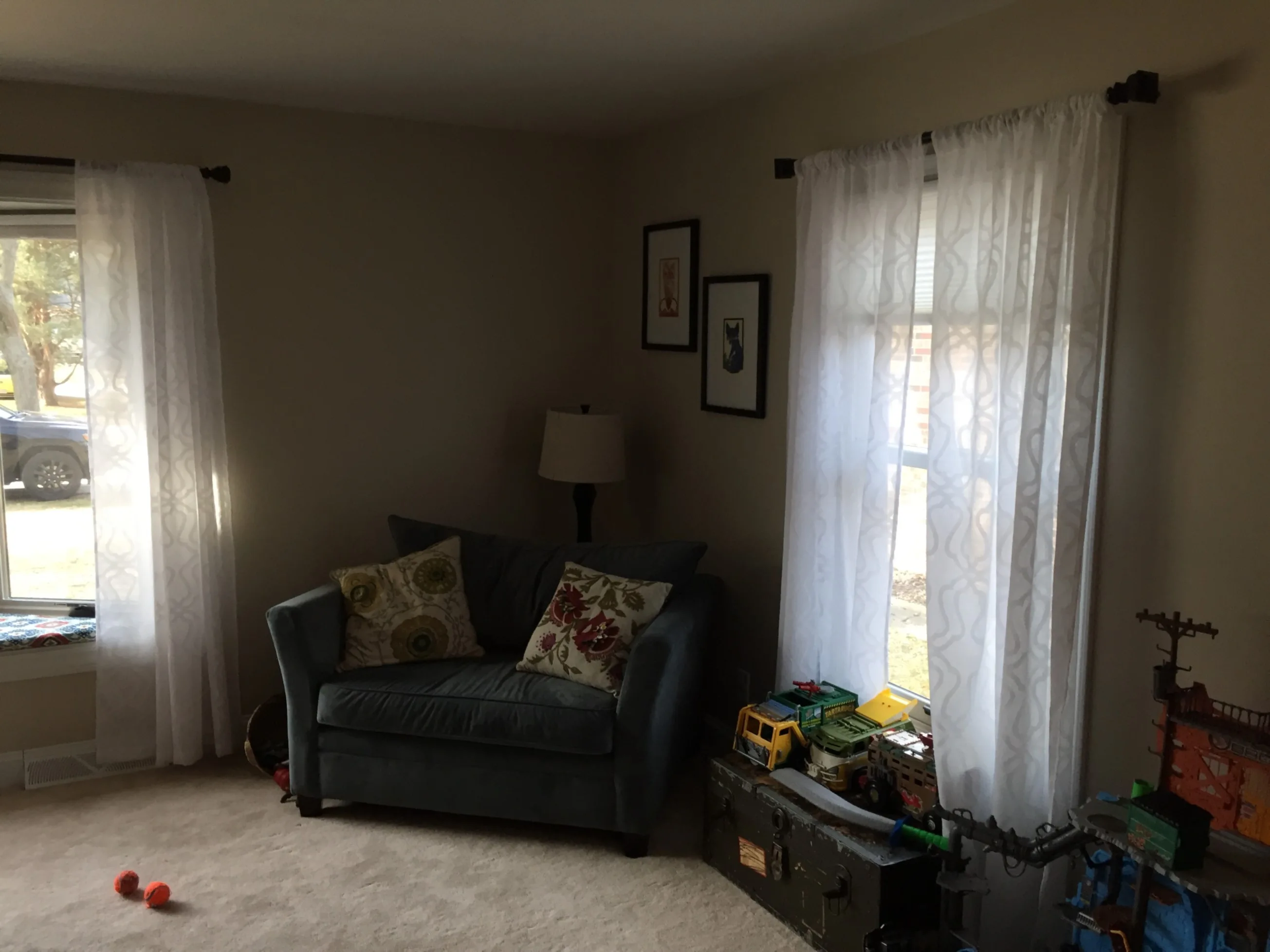 Cozy living room with gray couch, patterned cushions, white curtains, and toys by the window. Soft natural light illuminates the space.