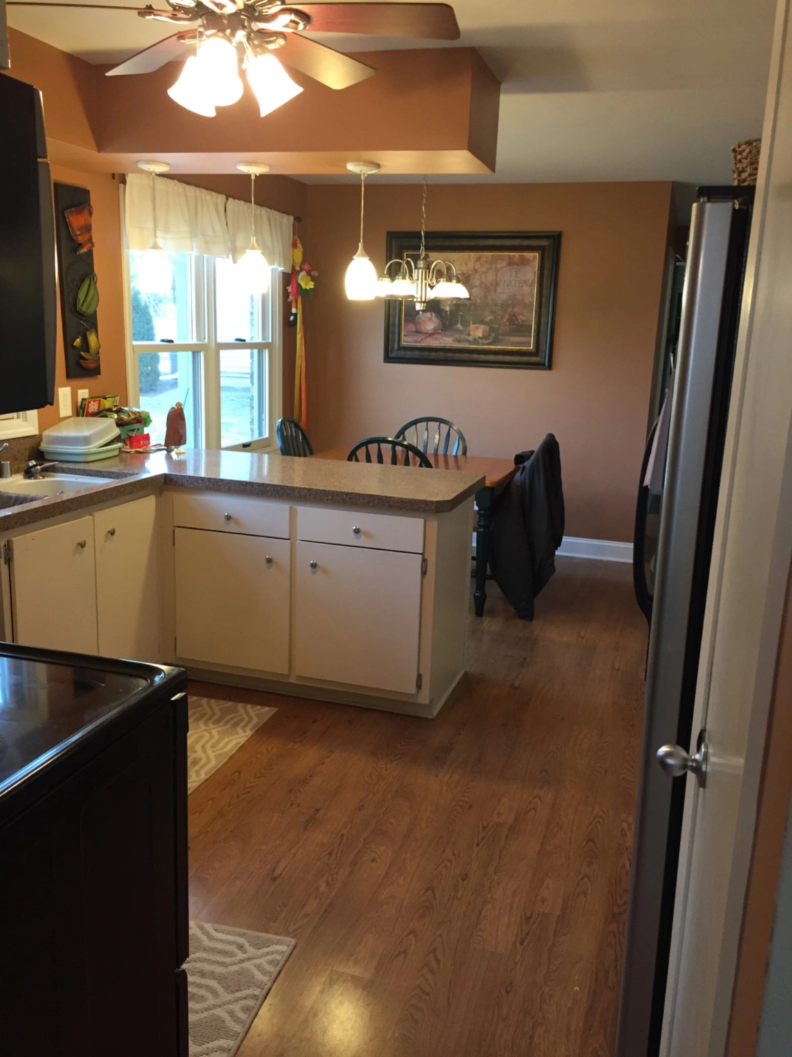 A cozy kitchen featuring wooden floors, ceiling fan, countertop with seating, and soft lighting, decorated with paintings and hanging lights.
