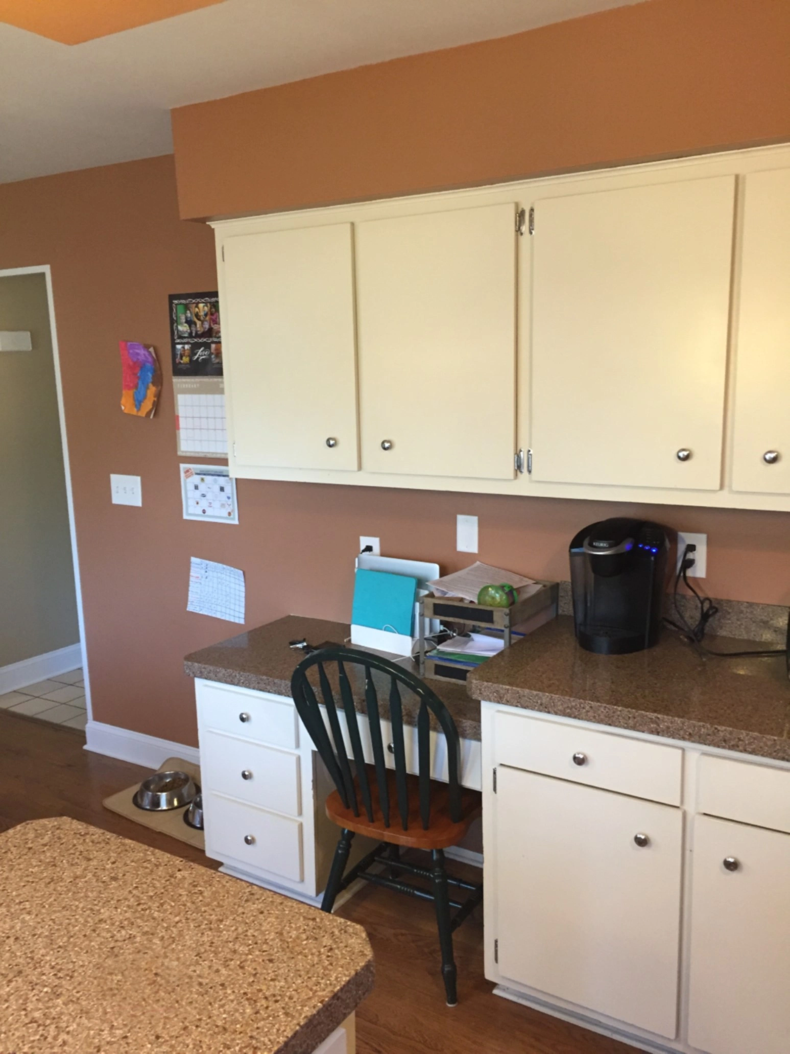 A home kitchen features beige cabinets, a small desk with a chair, colorful wall calendar, countertop items, and a coffee maker.