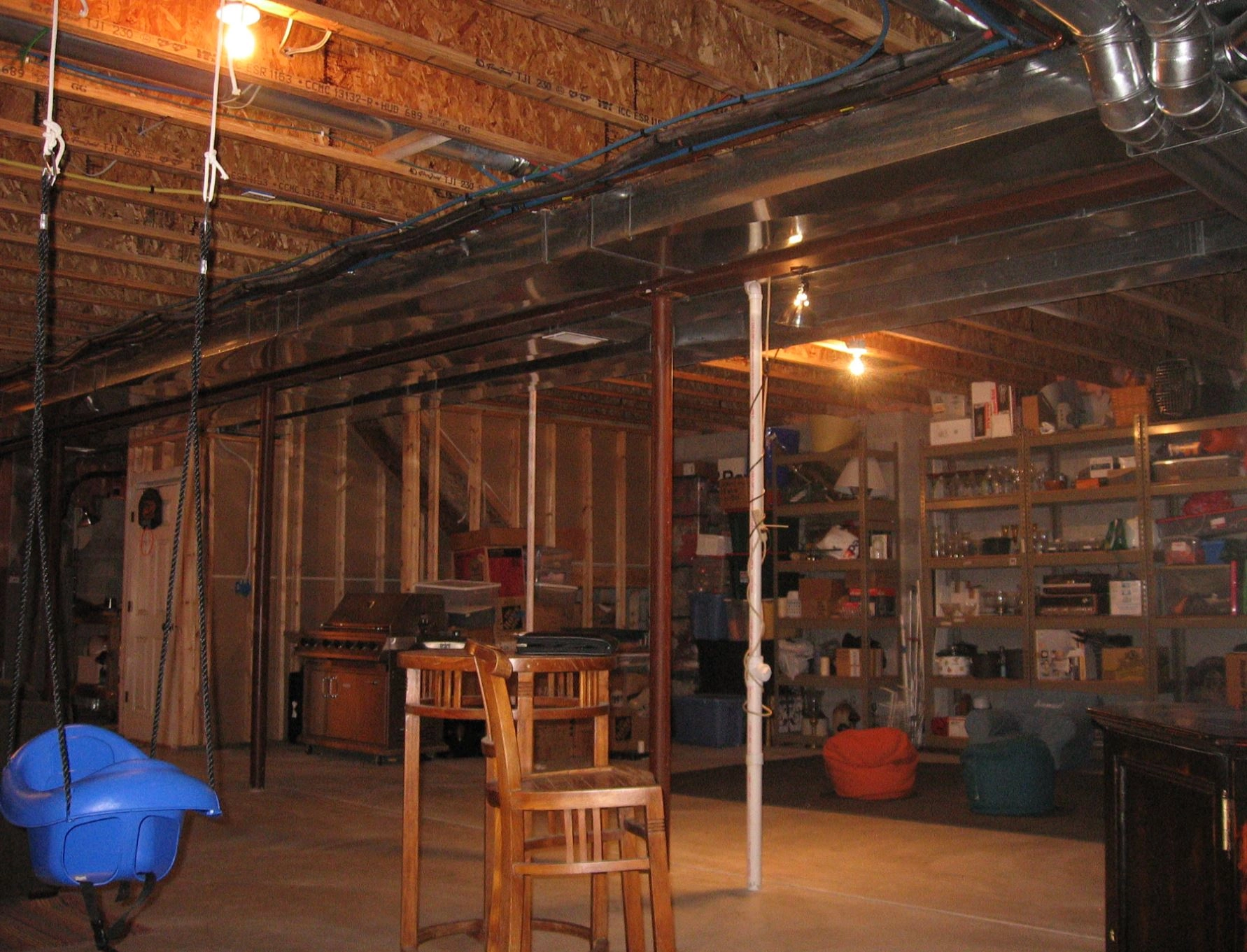 A basement with exposed beams, shelves filled with boxes, a swing, and wooden furniture under warm lighting. Equipment and storage are visible.