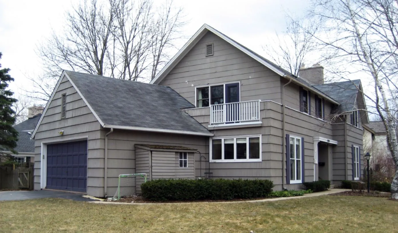 A two-story suburban house with gray siding, a blue garage door, and a small balcony, surrounded by bare trees and a lawn.