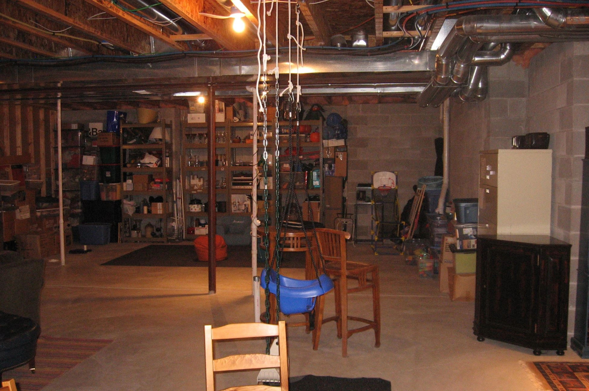 A dimly lit basement with exposed beams, shelves filled with boxes, a child's swing, chairs, and scattered storage items throughout the space.