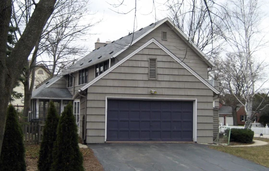 A suburban house with a gray exterior and two-car garage in a residential neighborhood lined with trees and nearby homes.