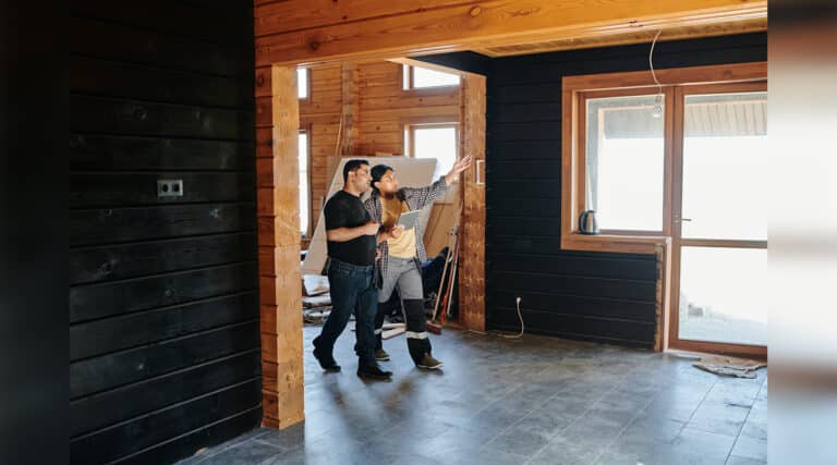 Two people discuss interior design in a wooden cabin under construction. One person gestures towards a window, while the other holds a tablet.