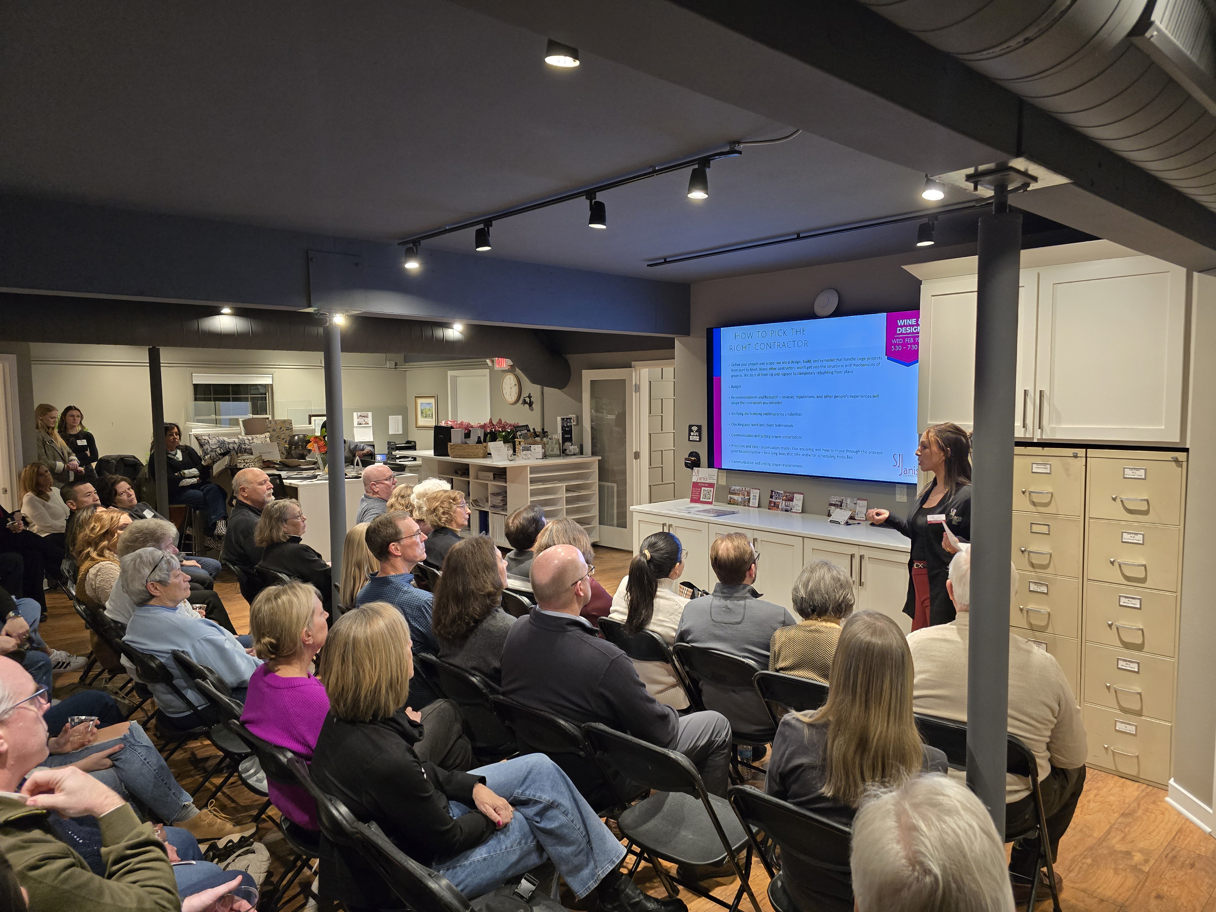 A person gives a presentation to a seated group in a modern room, featuring a screen displaying text and storage cabinets.