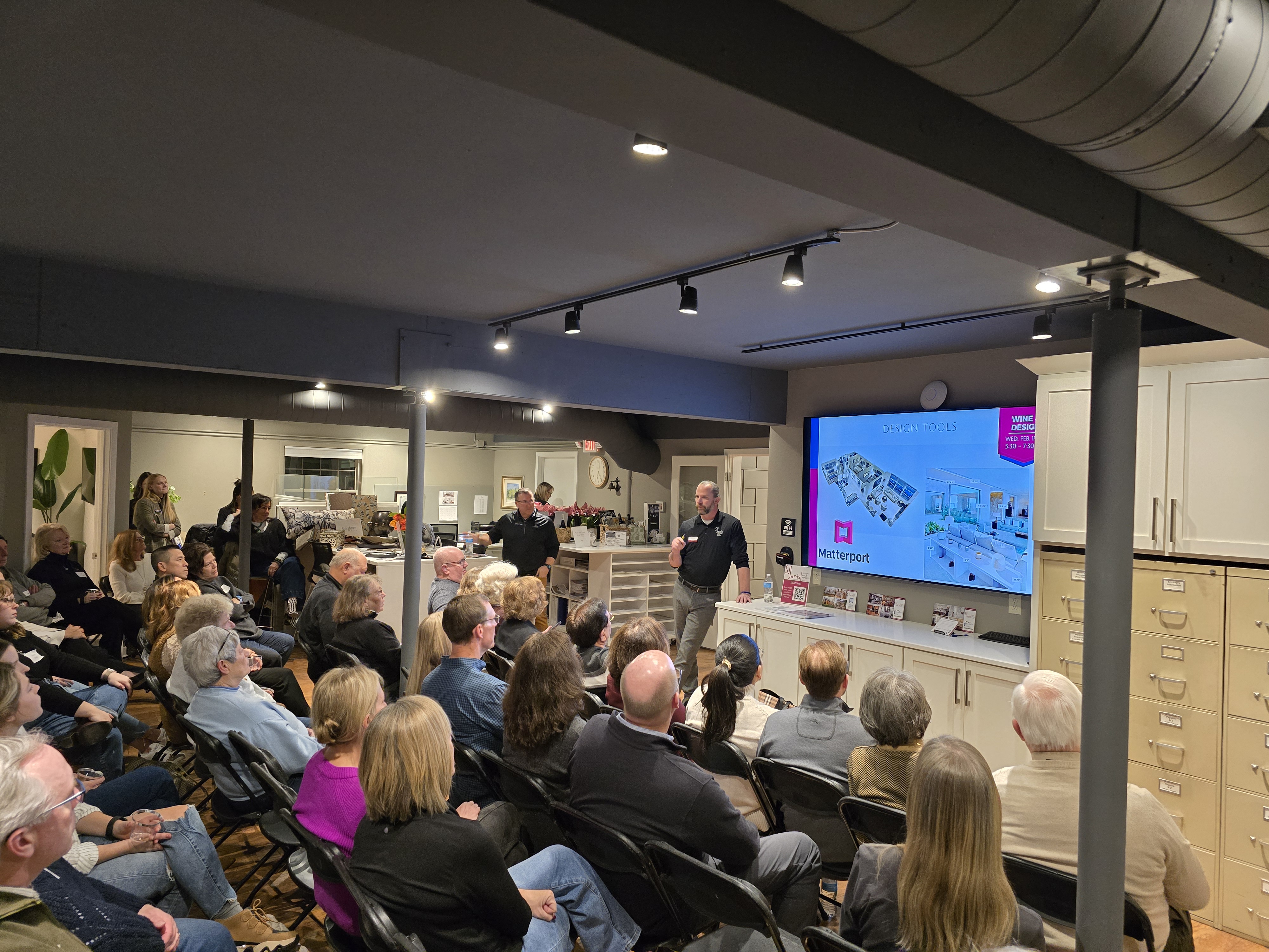 A group of people attentively listen to a presentation in a modern room, showcasing Matterport design tools on a screen.