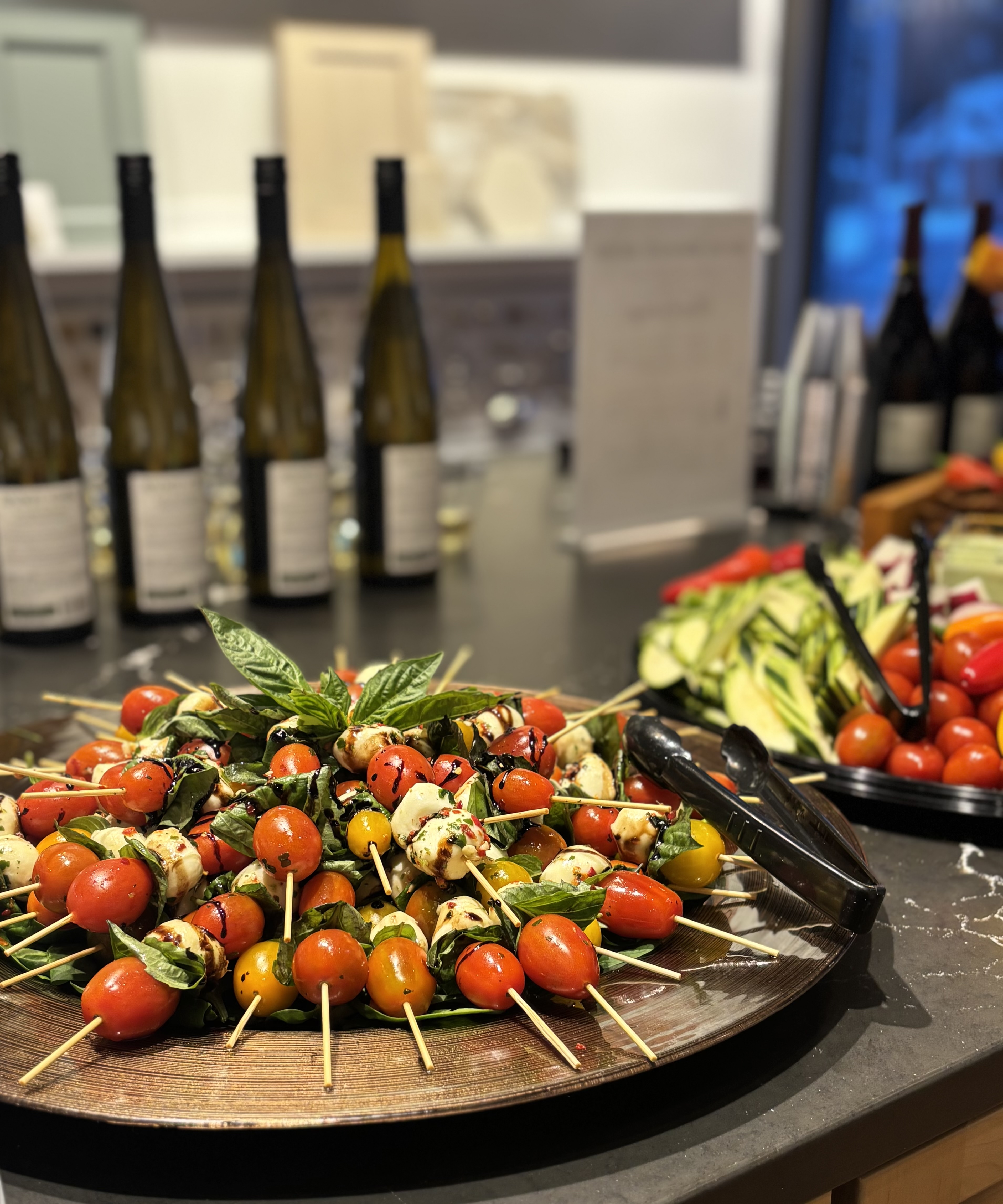 A platter of vibrant Caprese skewers, featuring cherry tomatoes and mozzarella, is displayed on a table alongside wine bottles and fresh vegetables.