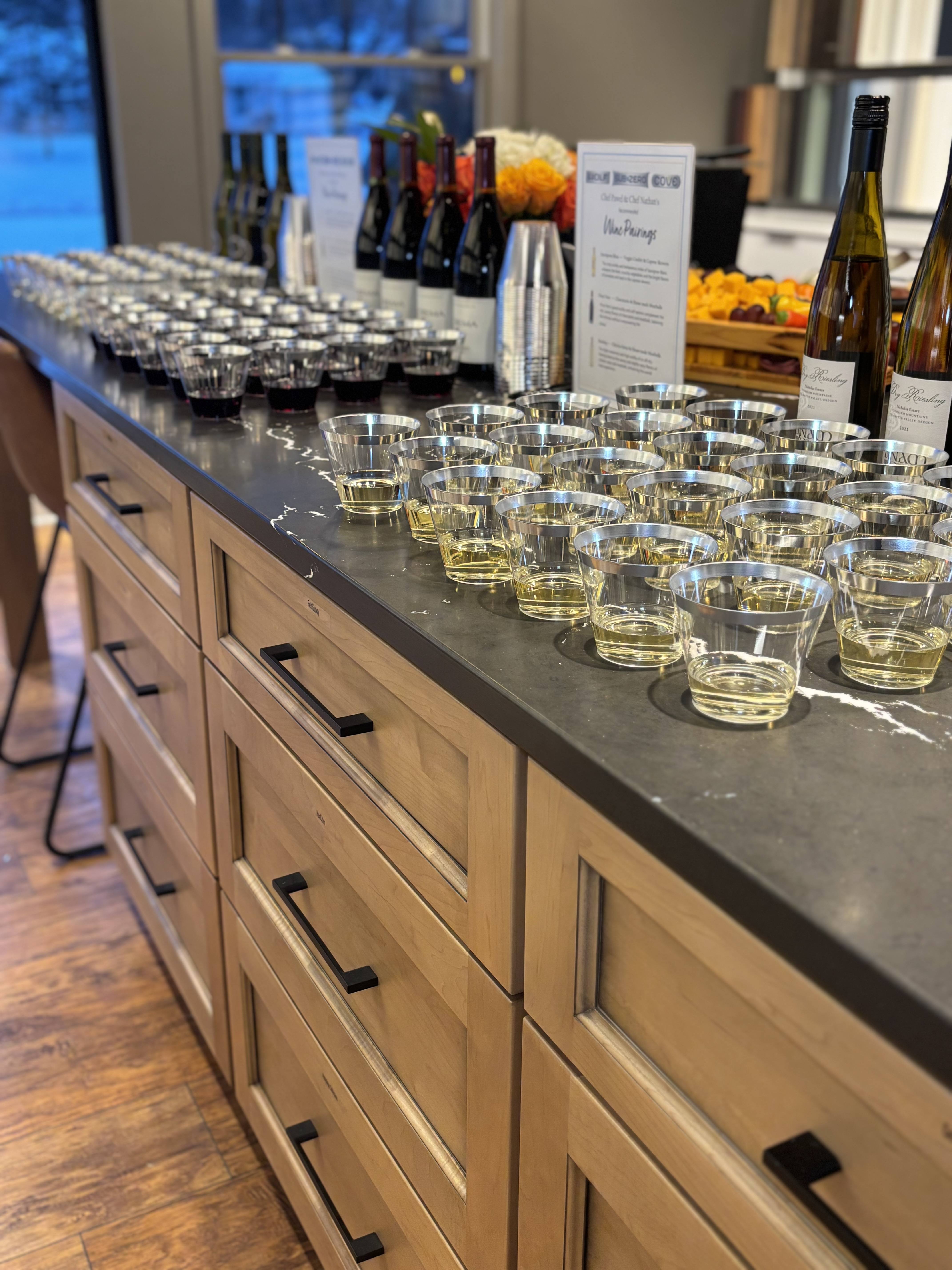 A counter displays rows of wine bottles and filled glasses, ready for tasting, with signs and floral decorations in the background.