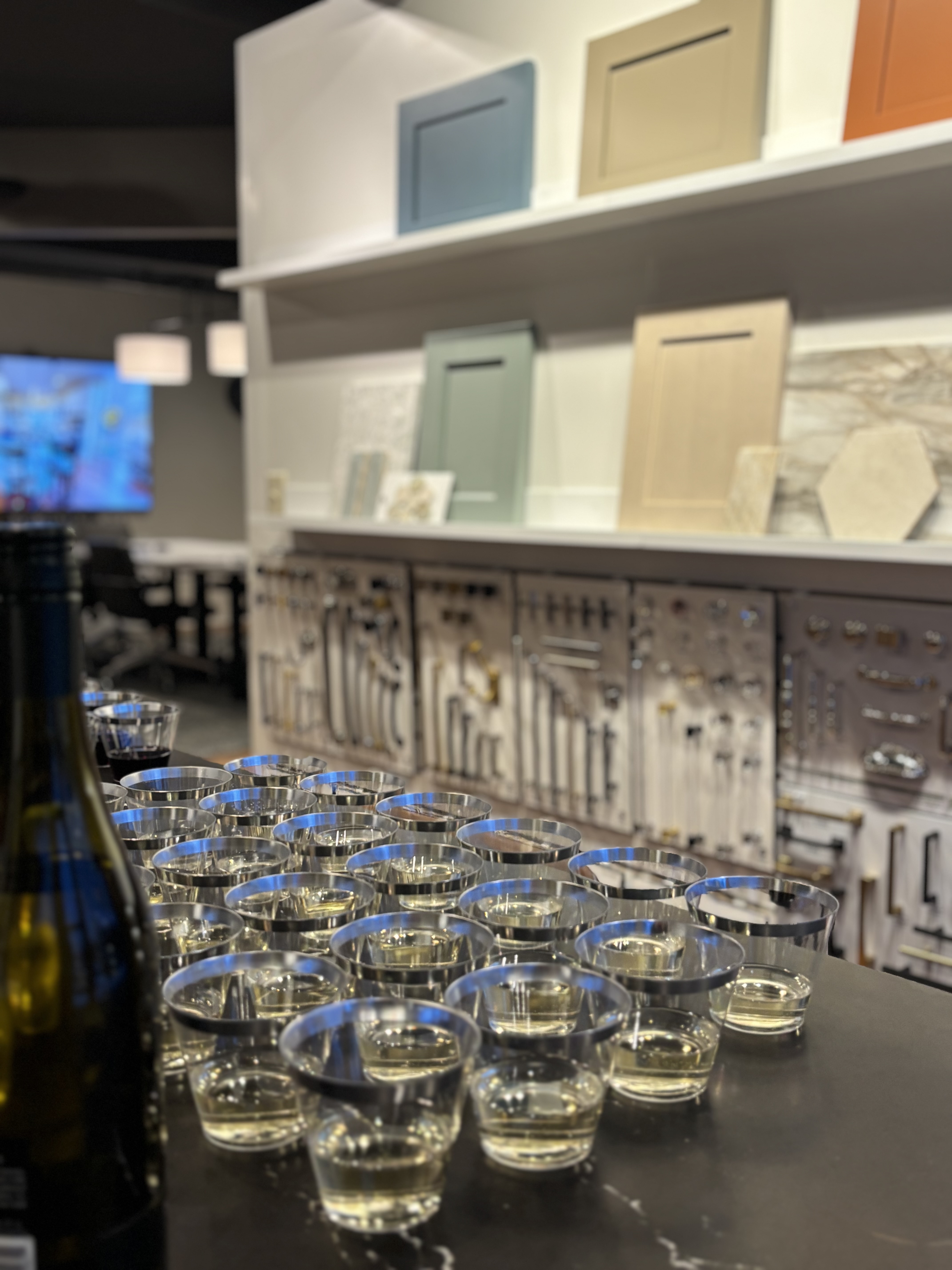 A countertop with multiple glasses of white wine and a bottle, set in a modern showroom displaying cabinet doors and hardware samples.