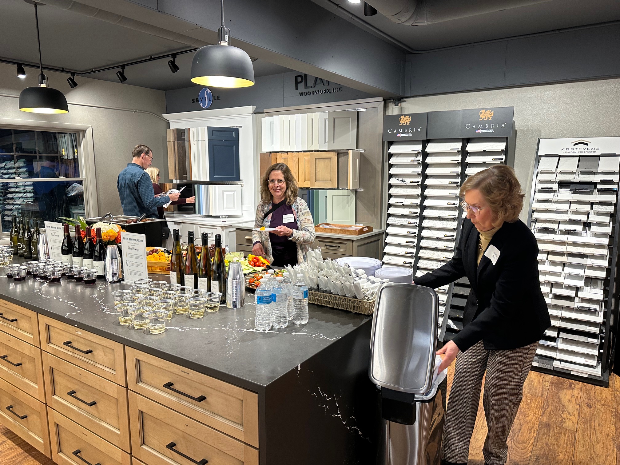 Three people interacting in a showroom with wine, glasses, and food on a counter. Cabinets and samples displayed in background.