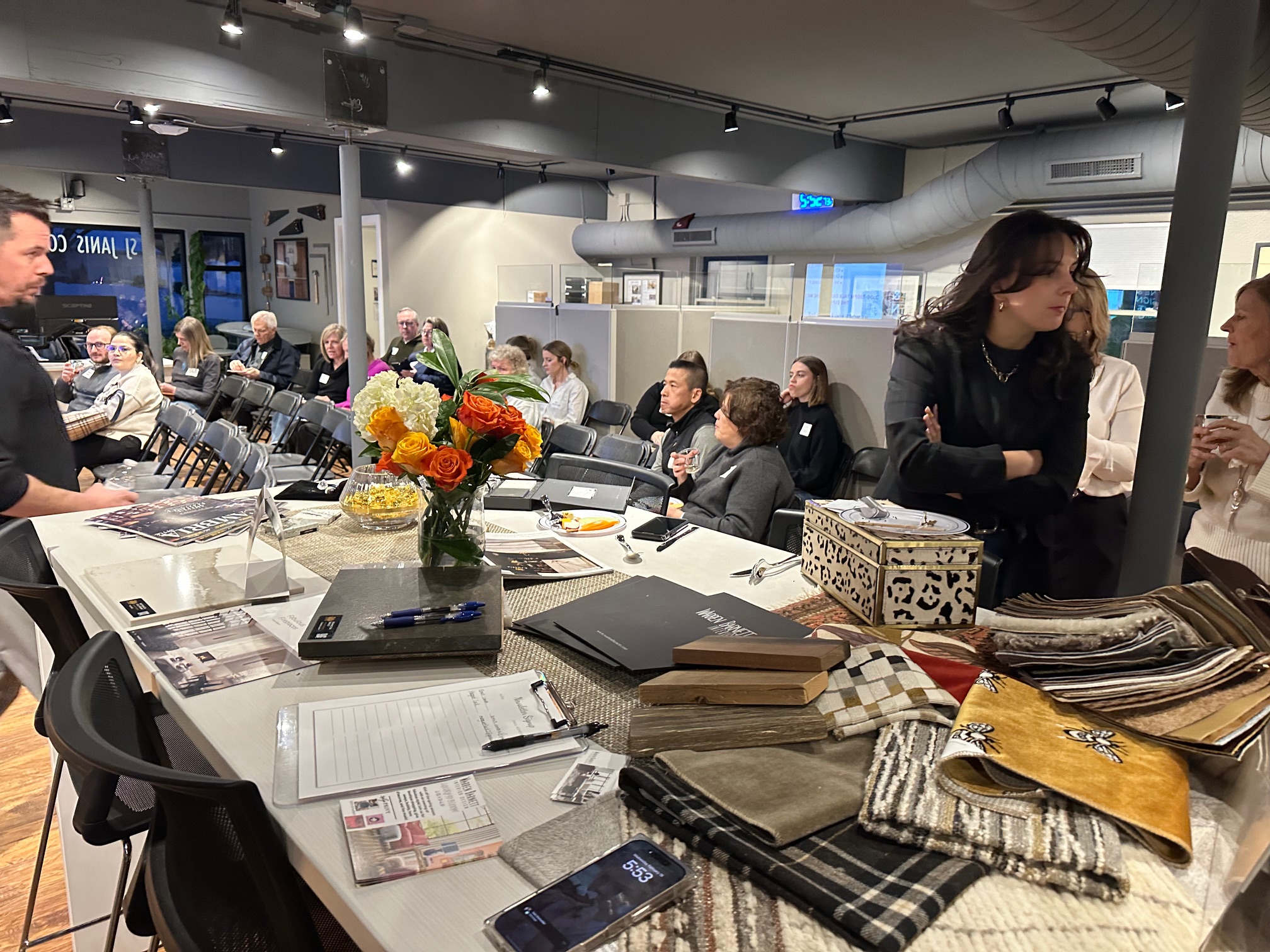 A group of people in a modern conference room surrounds a table with flowers and fabrics. Charts and pamphlets are visible.