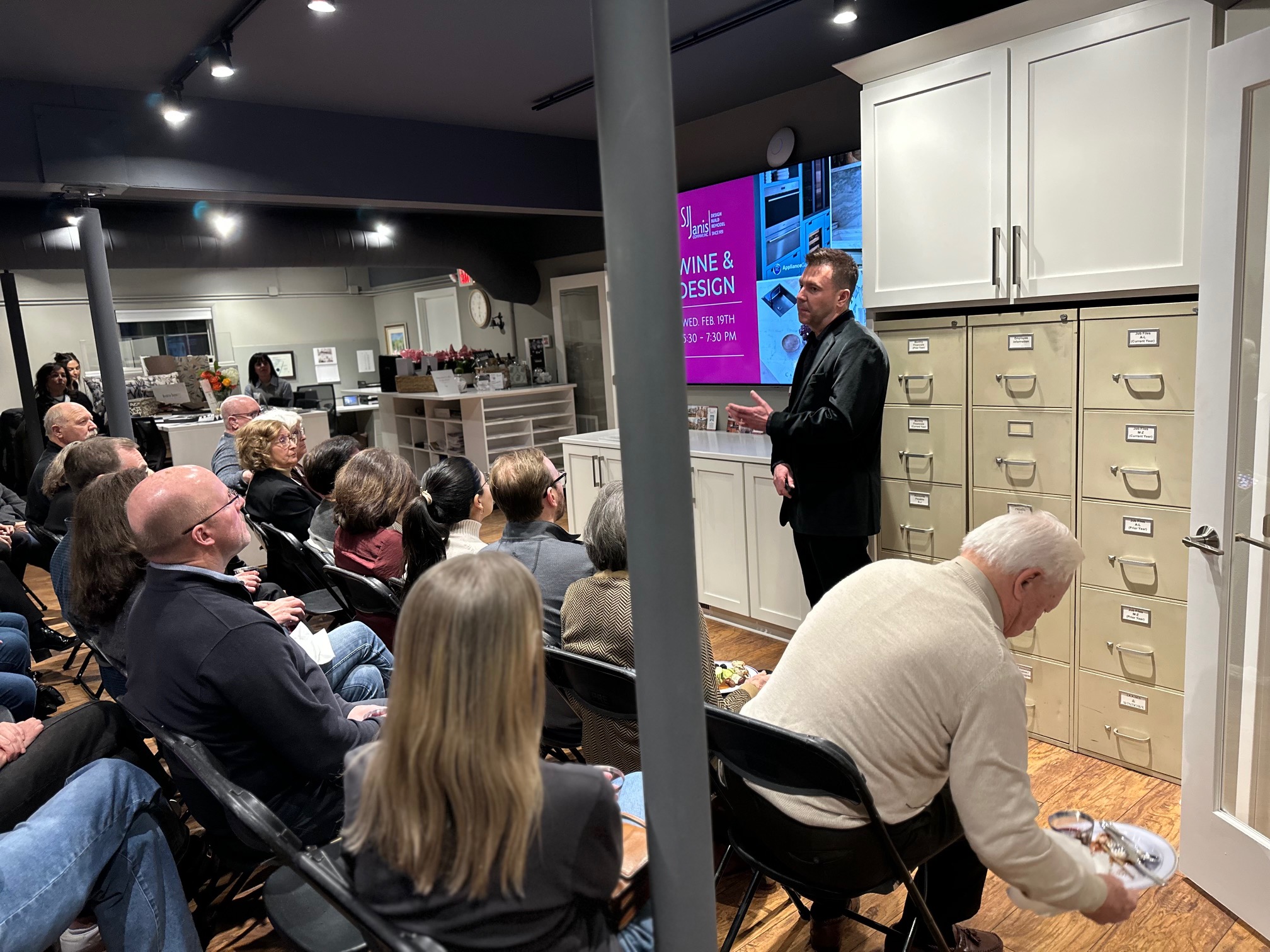 A person giving a presentation to an audience in a modern office with stacked filing cabinets and a digital screen displaying event details.