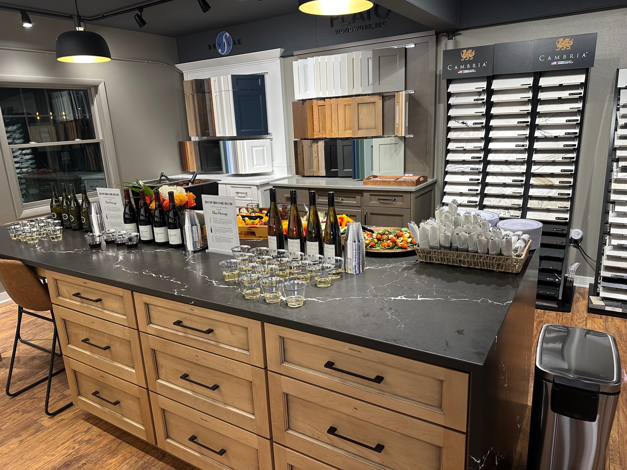 A kitchen showroom display featuring wine bottles, glasses, and appetizers on a marble counter; cabinetry and countertop samples are visible in the background.