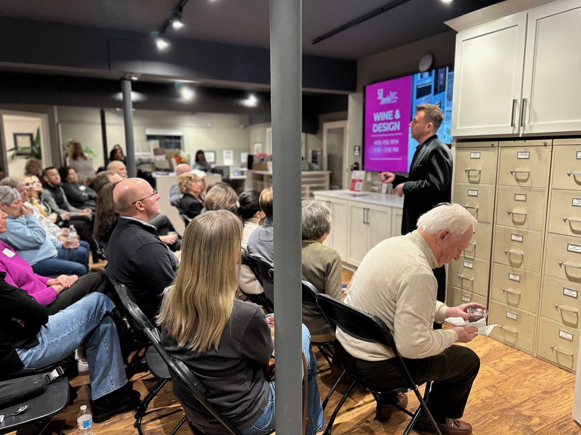 A group of people listen to a presentation in a room with a "Wine & Design" sign, featuring rows of filing cabinets.