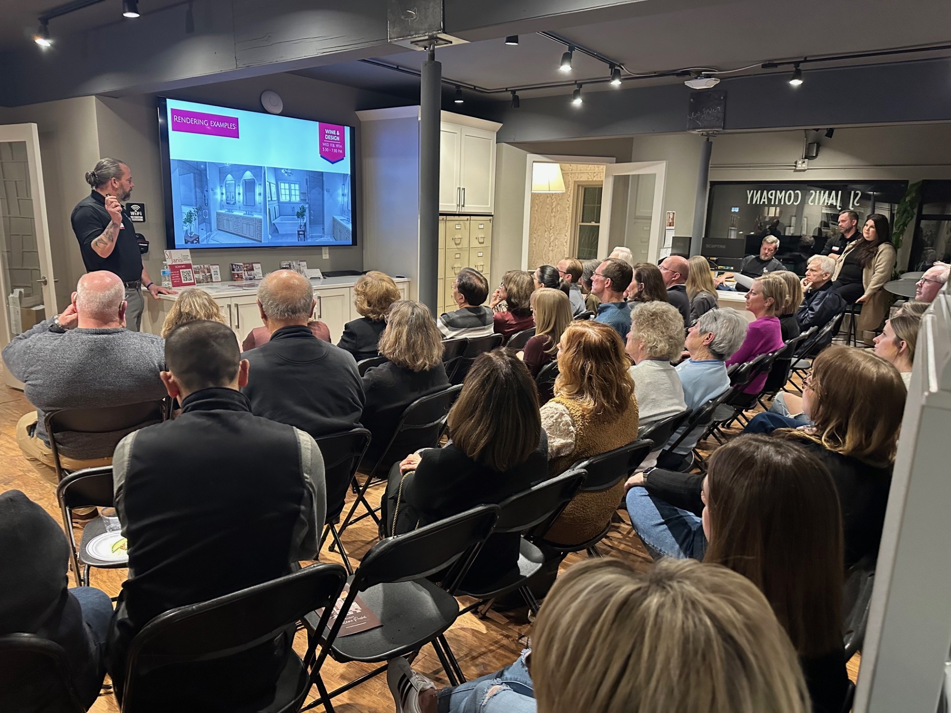 A person presents interior design visuals to a seated audience in a modern room with a large screen display.