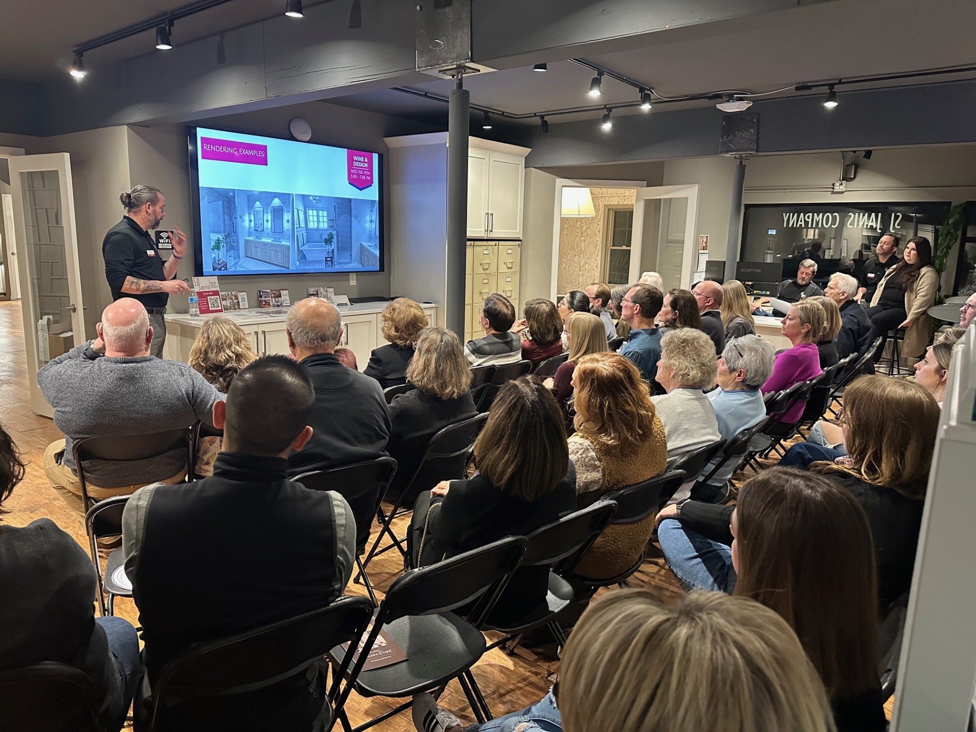 A person presents interior design renderings to an attentive audience in a modern showroom setting, featuring bright lighting and decor.