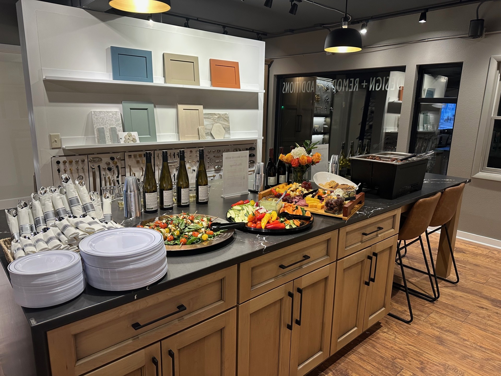 A stylish kitchen showroom features a countertop with a spread of food, wine bottles, and neatly arranged plates and napkins.