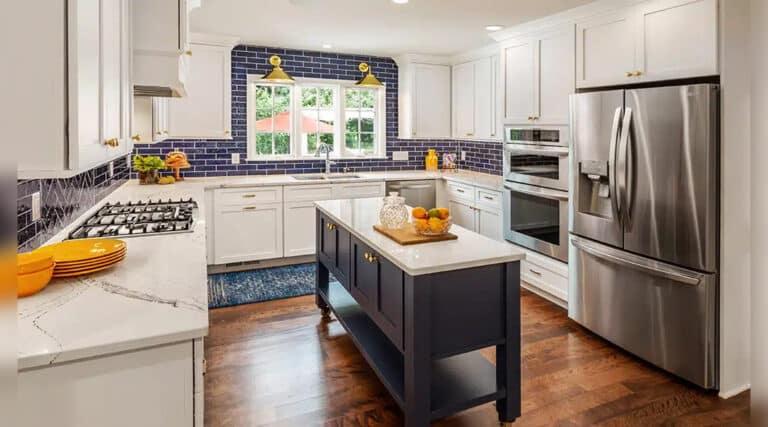 A modern kitchen features white cabinets, stainless steel appliances, a navy island, and blue subway tile backsplash with natural light through a large window.