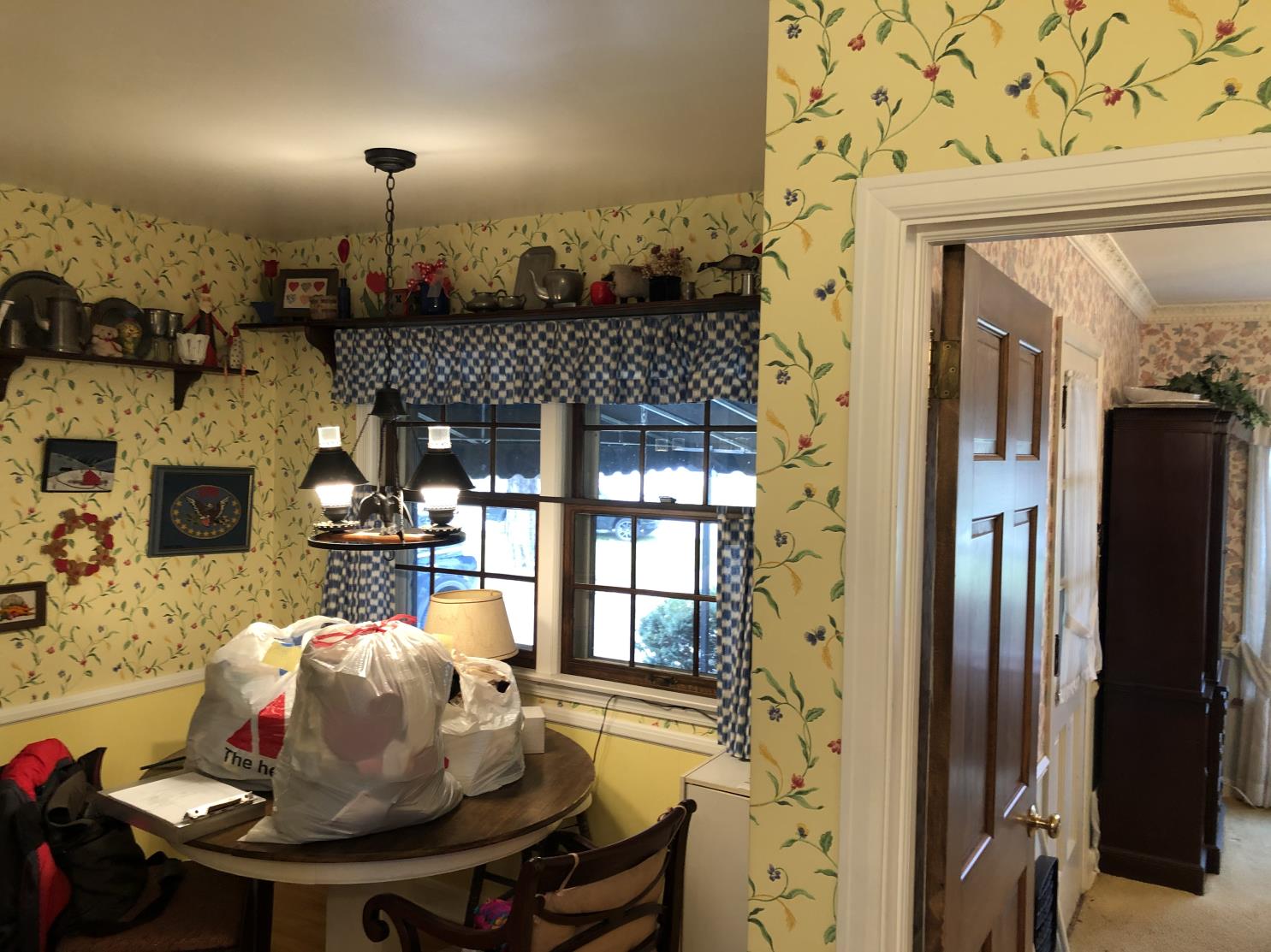 A cozy kitchen with floral wallpaper, a round table cluttered with bags, and shelves showcasing decorative teapots and cans under a hanging light.