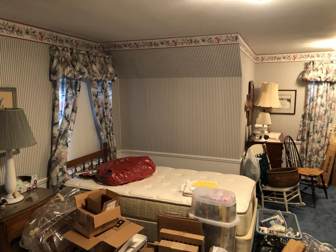 A cluttered bedroom with floral curtains, wallpaper, and various items scattered on the bed and floor, including boxes and lamps.