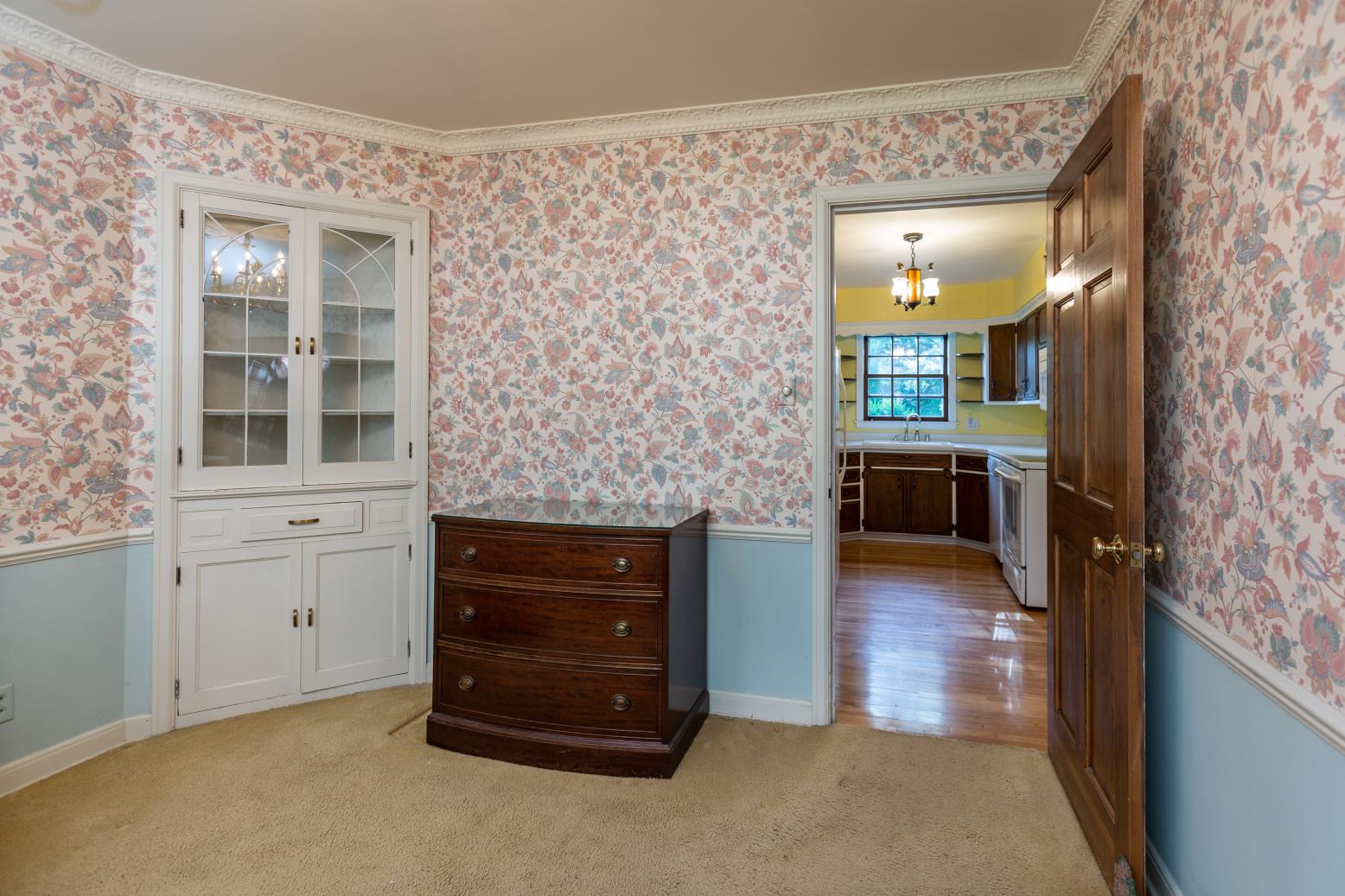 A cozy vintage room features floral wallpaper, a wooden dresser, and china cabinet. Doorway opens to a bright kitchen with wooden cabinets.