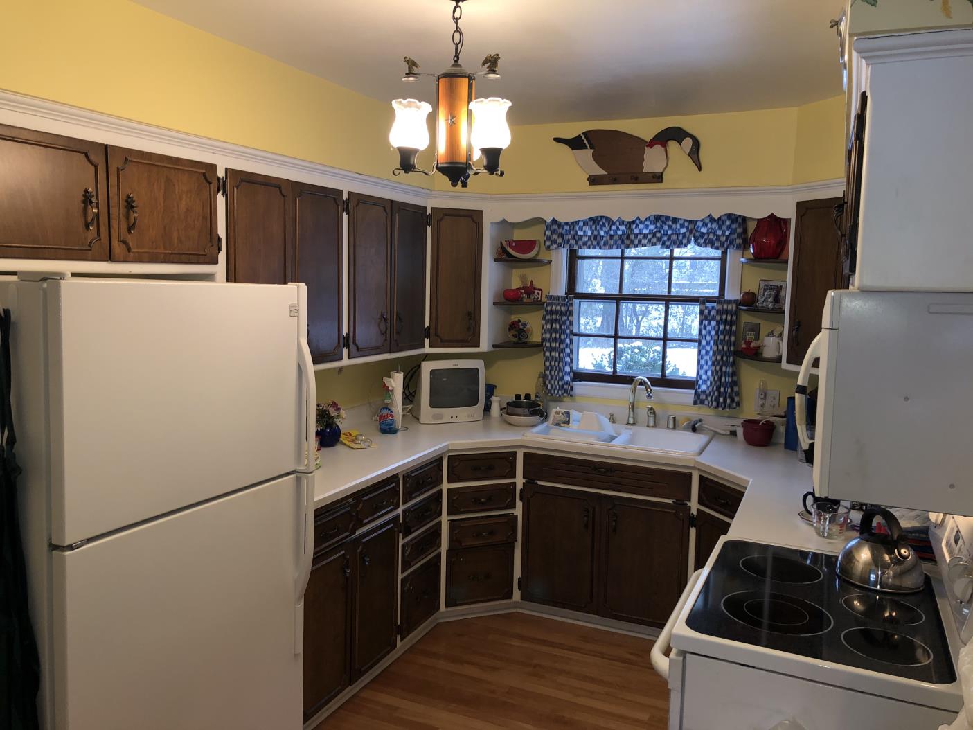 A kitchen with yellow walls, wooden cabinets, and a white countertop. It features a refrigerator, stove, vintage TV, and duck wall decor.