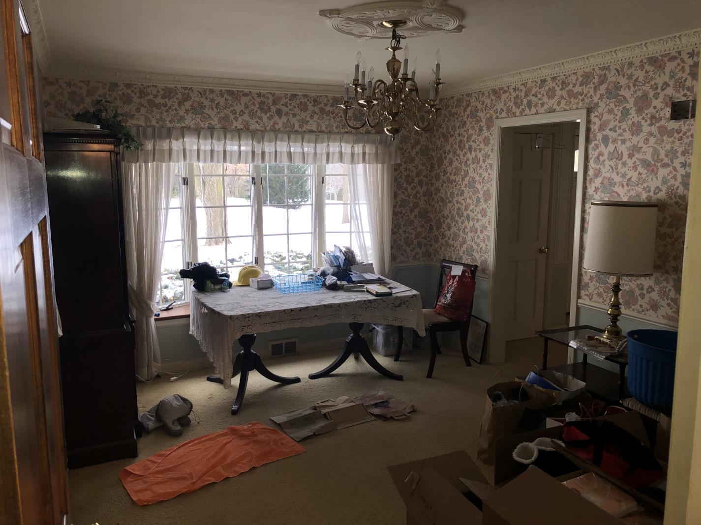 A cluttered room with floral wallpaper, chandelier, and large window. A table is covered with household items; boxes and bags on the floor.