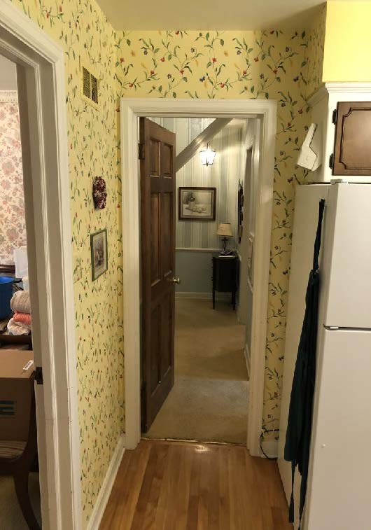 A narrow hallway features floral wallpaper, leading to a carpeted room with artwork and a lamp on a small table.
