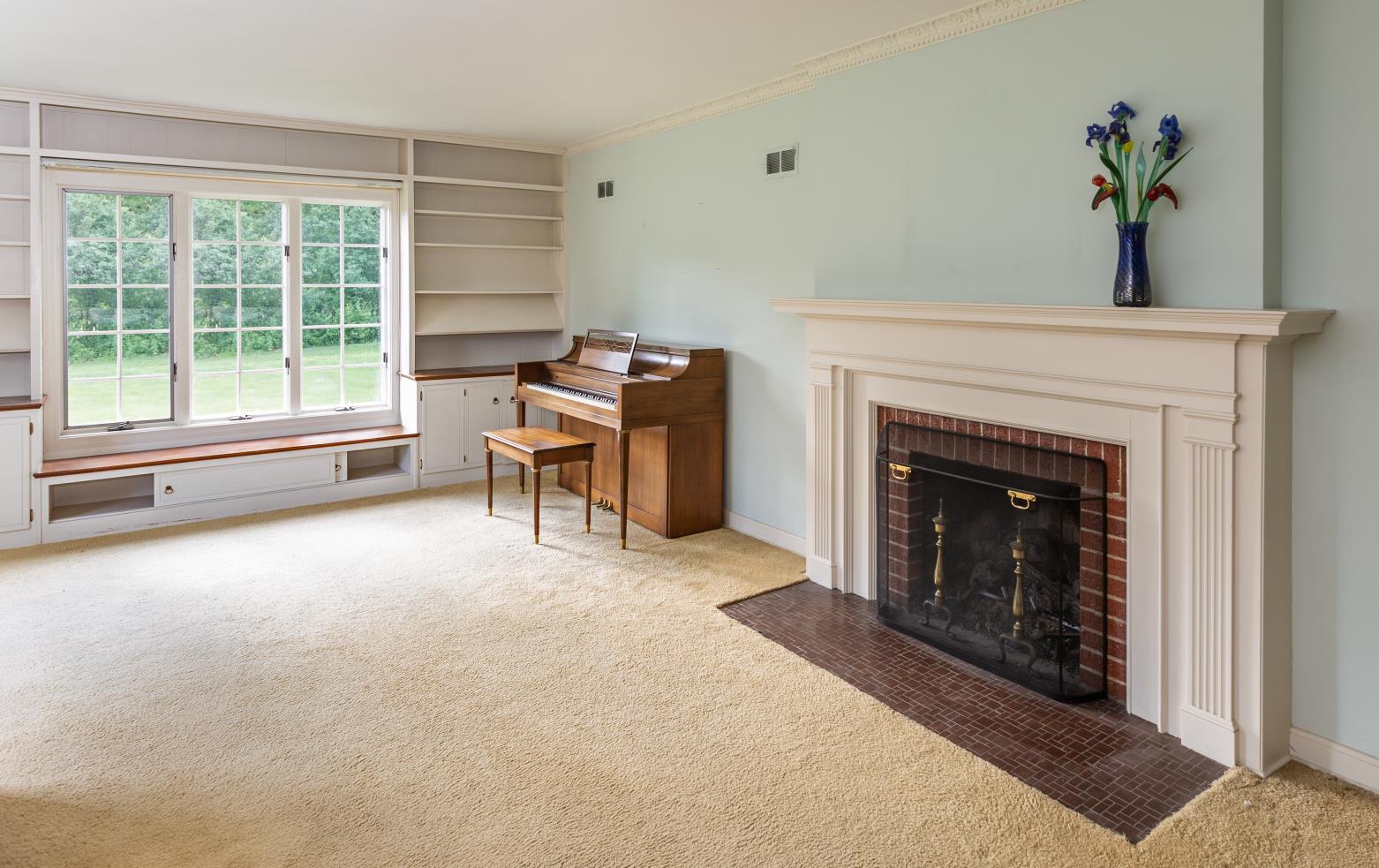 A cozy living room features a piano, fireplace, large window with garden view, and built-in shelves. A vase with flowers adds decor.