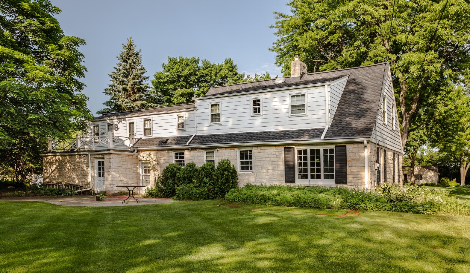 A charming two-story house with a Mansard roof, surrounded by lush greenery and trees, featuring large windows and a sunny front yard.