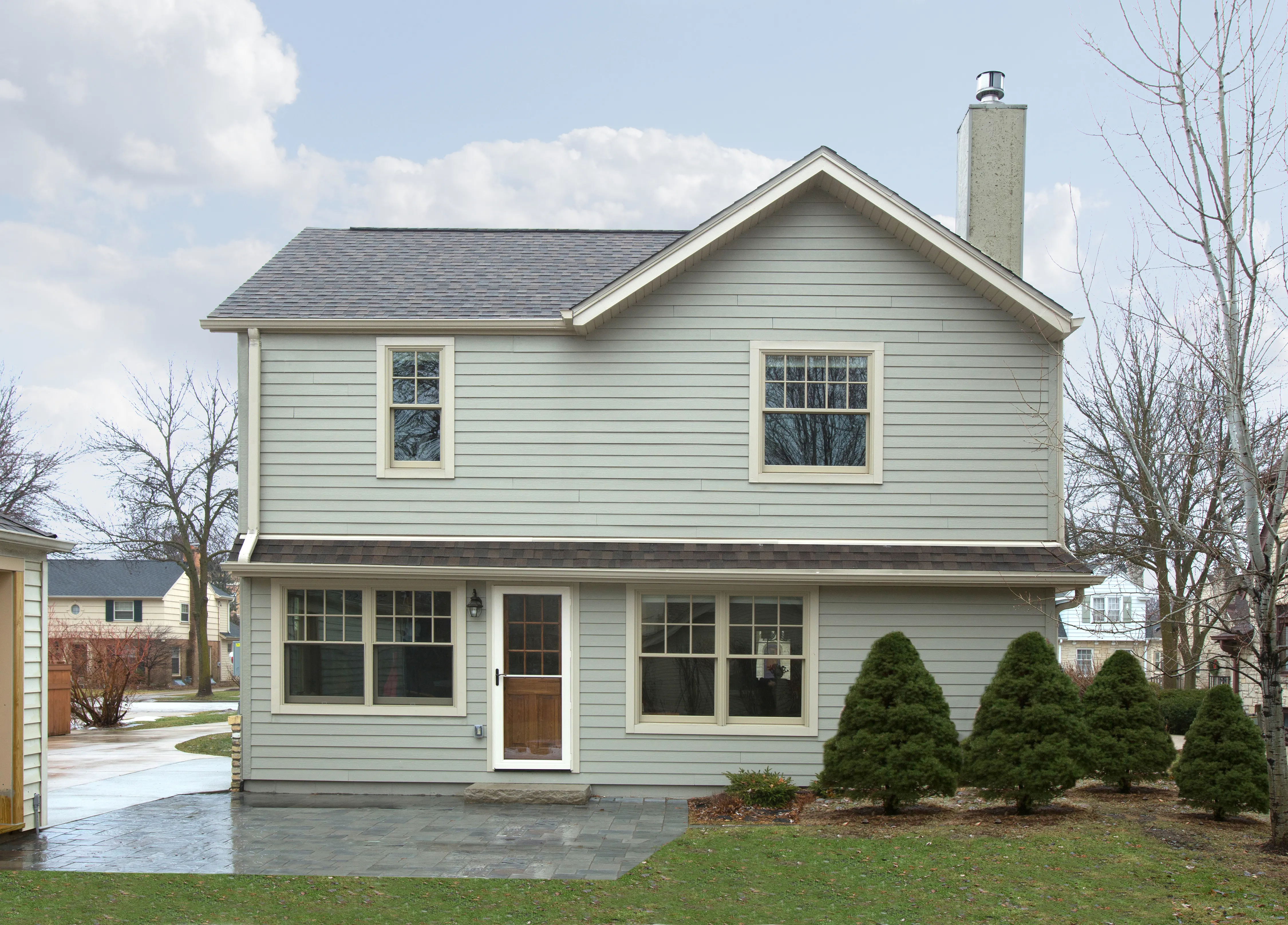 A two-story gray house with a chimney, surrounded by bushes and trees on a clear day, with neighboring houses in view.