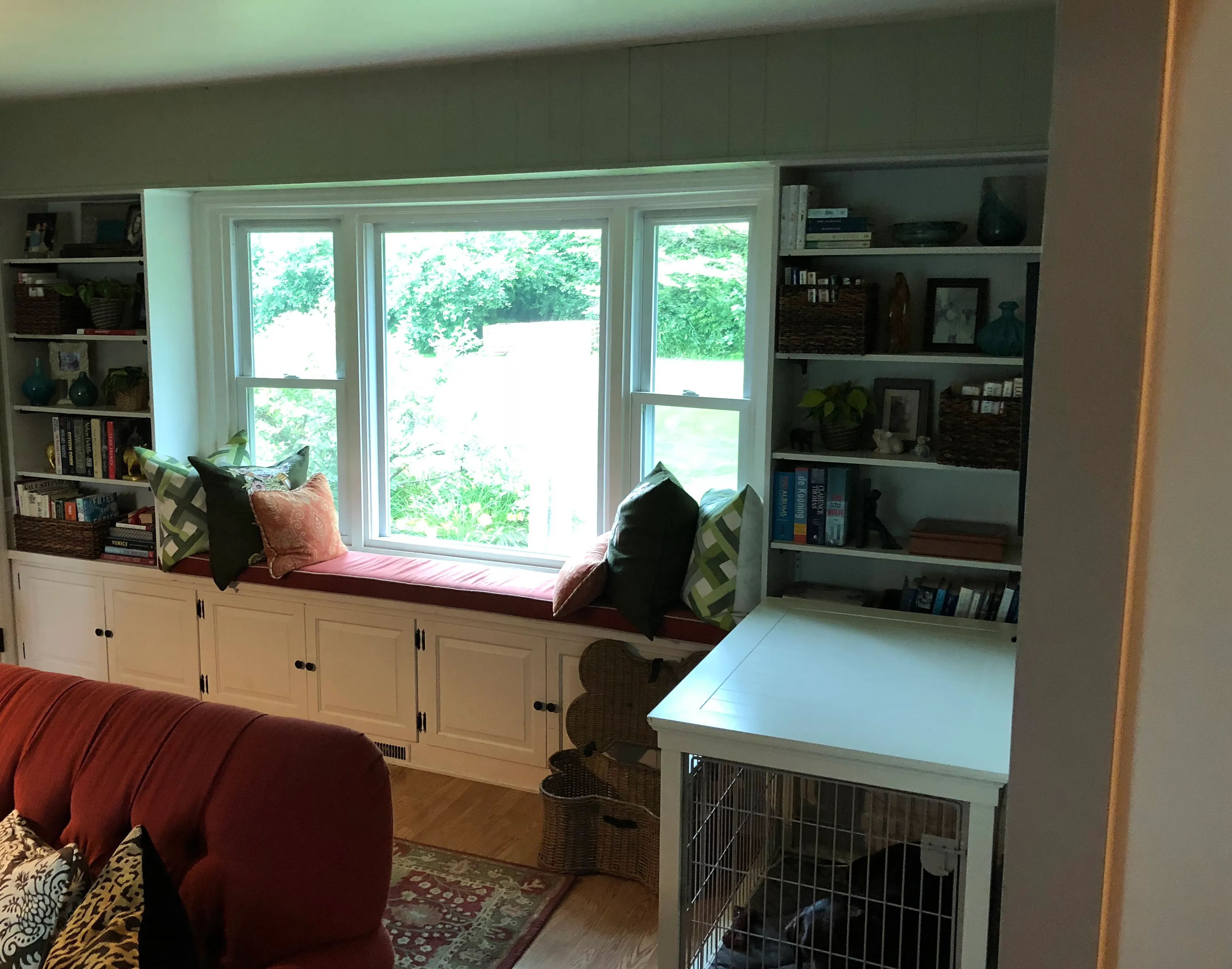 Cozy living room with built-in shelves, bay window seat, colorful cushions, and pet crate. Books and decor items adorn shelves.