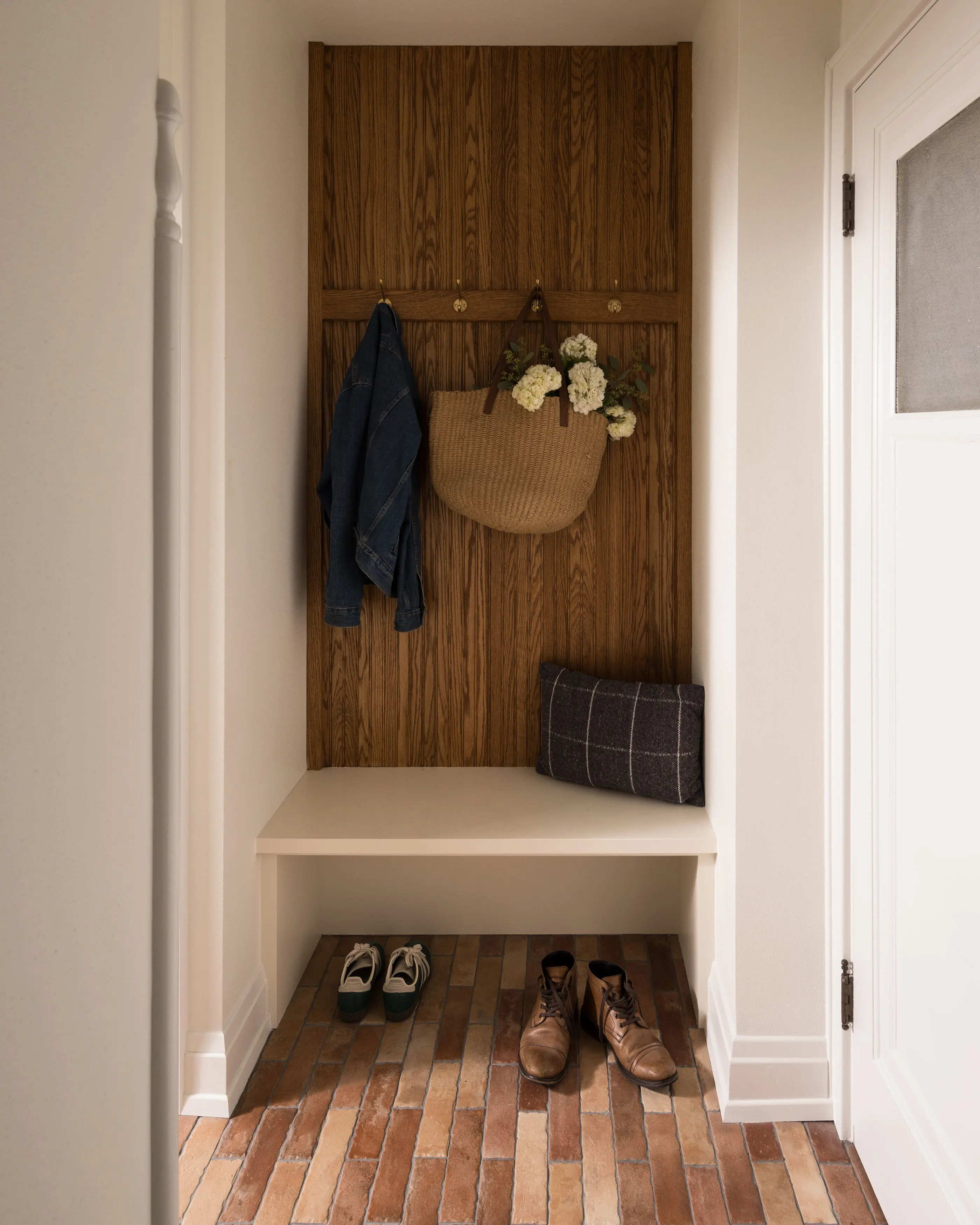 A cozy entryway features a wooden wall with hooks, holding a denim jacket and woven bag. Brick flooring displays shoes and a pillow.