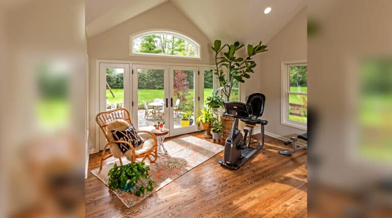 Bright sunroom with wooden floors, exercise bike, wicker chair, and potted plants; large windows overlook a green garden with patio seating.
