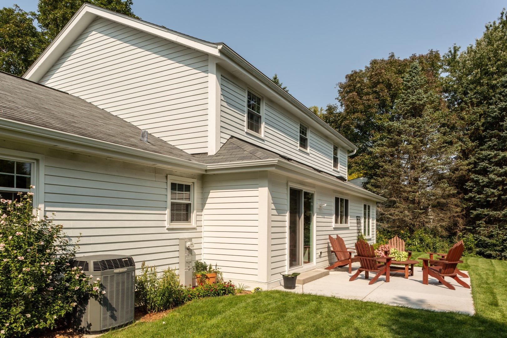 A suburban home with white siding features a backyard patio, Adirondack chairs, and lush green lawn surrounded by mature trees.