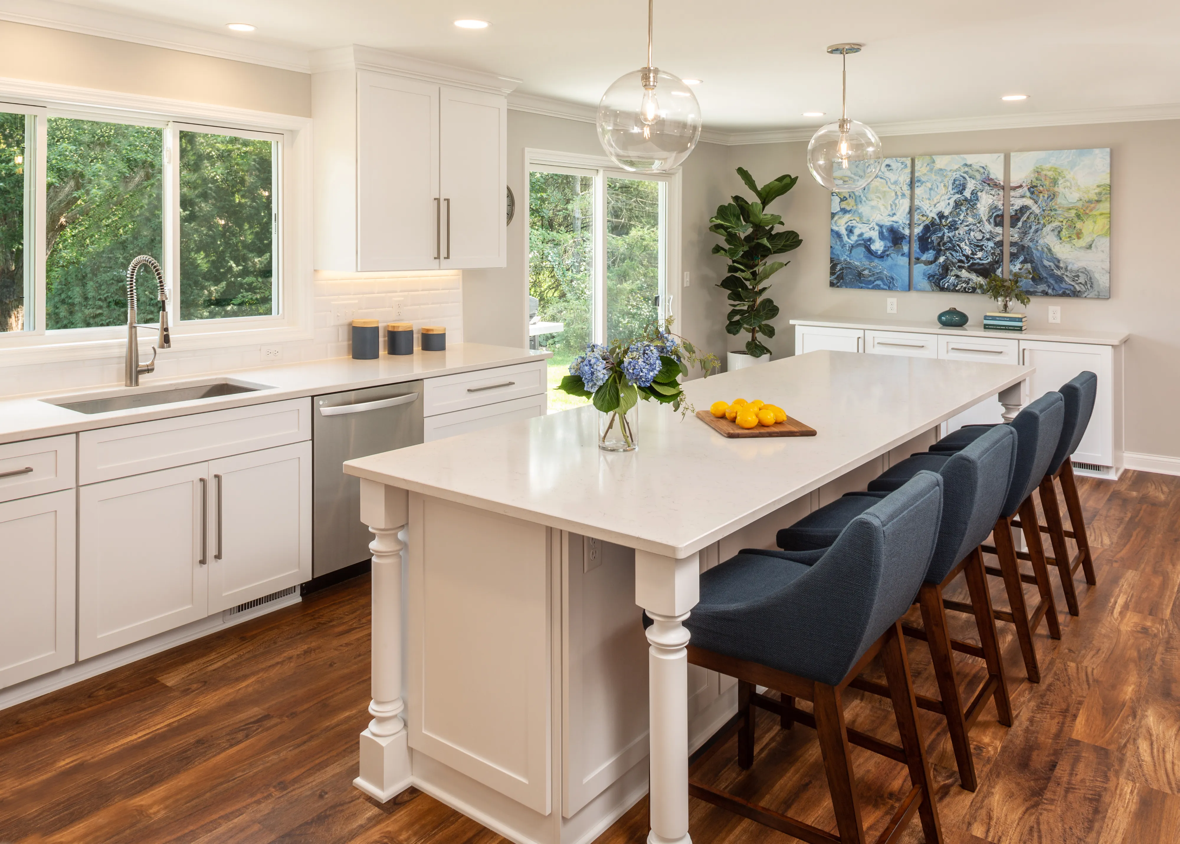 Bright kitchen with large island, blue chairs, flowers, and lemons. Wooden floor, modern décor, and large window overlooking green trees.