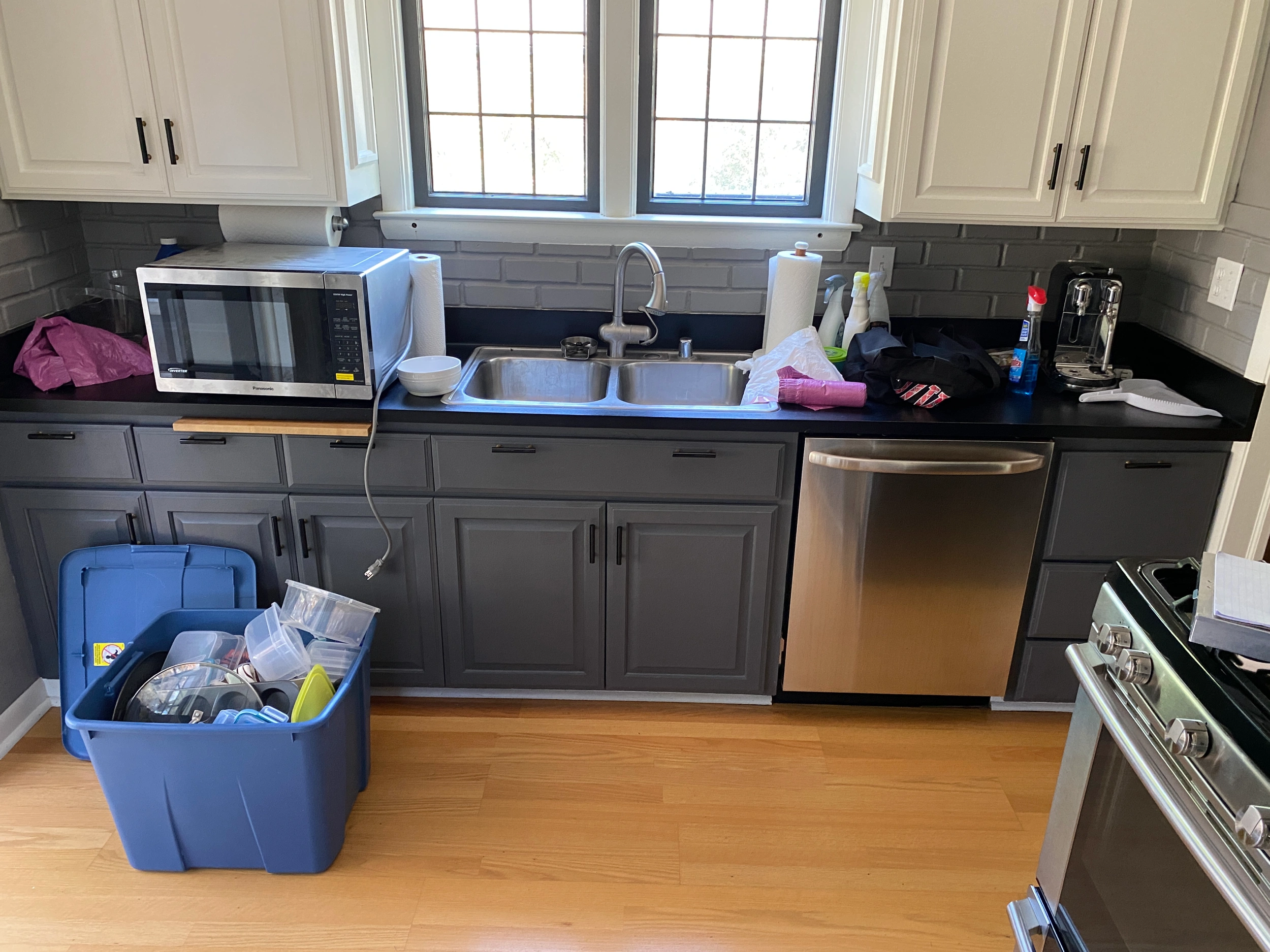 A kitchen displays gray cabinets, a microwave, sink, dishwasher, cleaning supplies, and open storage bin with containers on wooden floor.